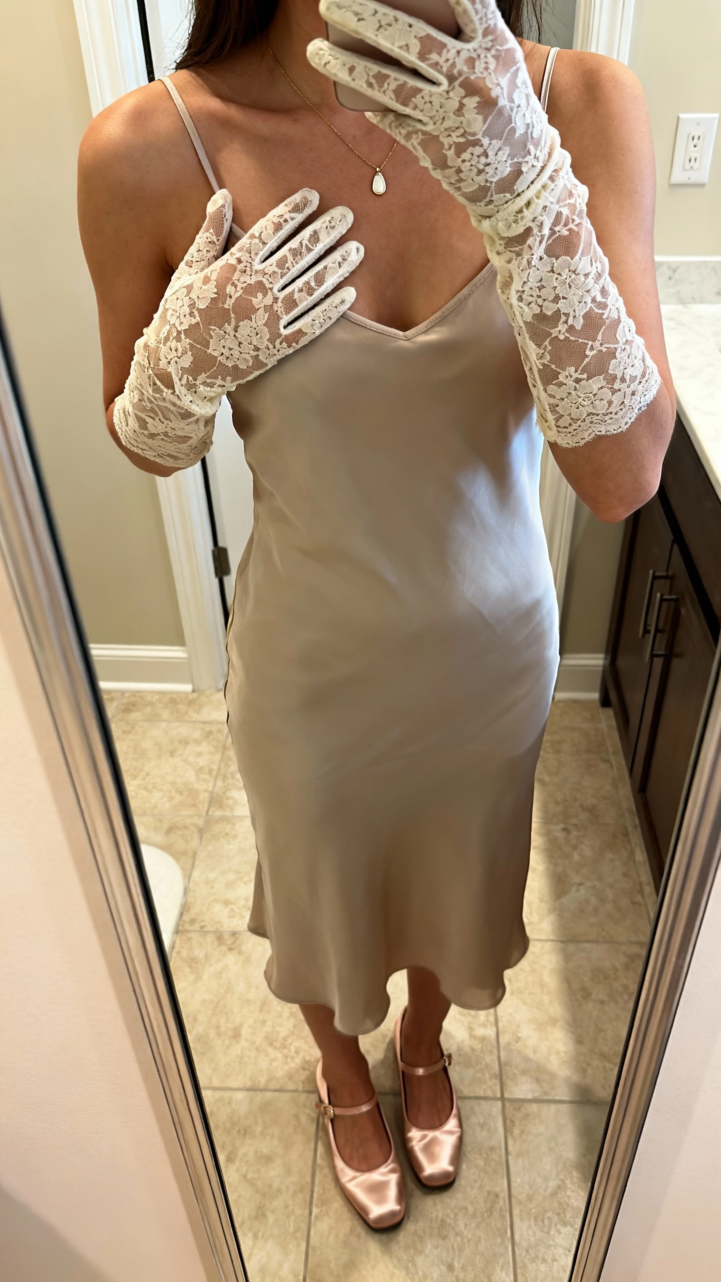 iPhone mirror selfie cropped at shoulders showing a woman wearing vintage-inspired lace opera gloves with a modern slip dress, satin mary janes, and a simple pendant necklace, face not visible, bathroom mirror with warm indoor lighting, natural iPhone photo quality.