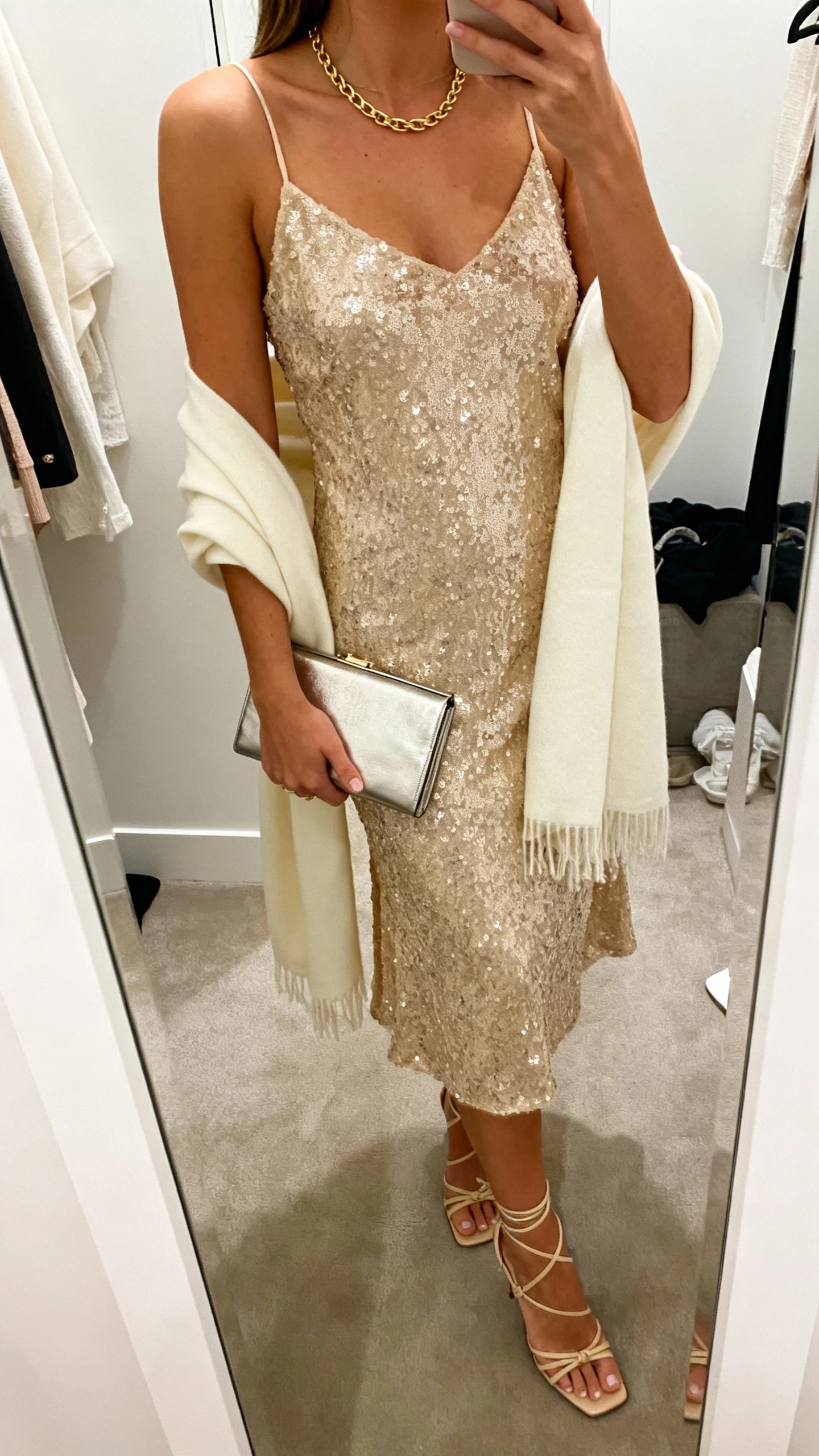 iPhone mirror selfie cropped at shoulders showing a woman wearing a champagne-gold sequin slip dress with a minimal gold chain necklace, strappy heels, a cream shawl, and a slim metallic clutch, face not visible, softly lit dressing room, natural iPhone aesthetic.