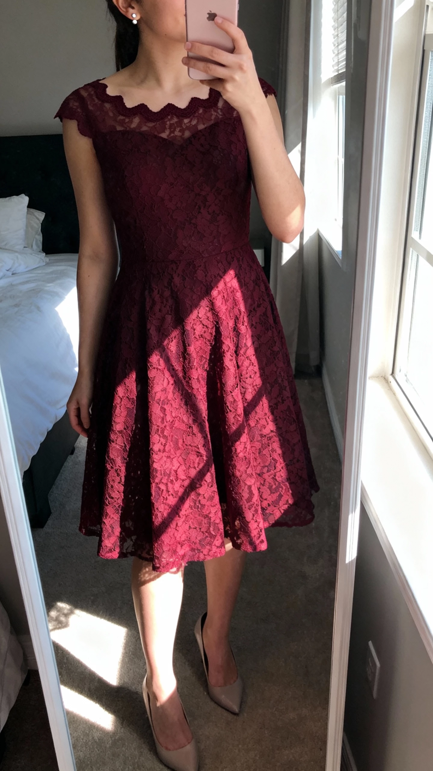 iPhone mirror selfie cropped at shoulders showing a woman in a burgundy lace A-line gown with a scalloped neckline, delicate stud earrings, classic pumps, face not visible, bedroom mirror, natural window light, iPhone photo quality.