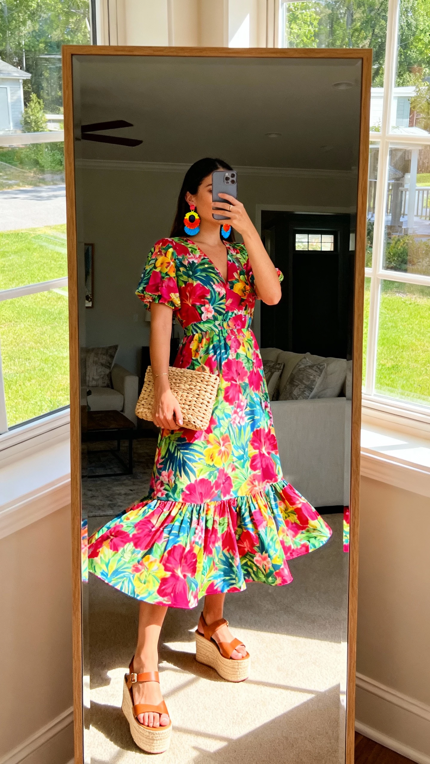 iPhone mirror selfie of a woman in a bright botanical maxi dress with twirl-friendly skirt, platform sandals, colorful statement earrings, and a woven clutch, face not visible, living-room full-length mirror, sunny window light, natural iPhone photo quality.