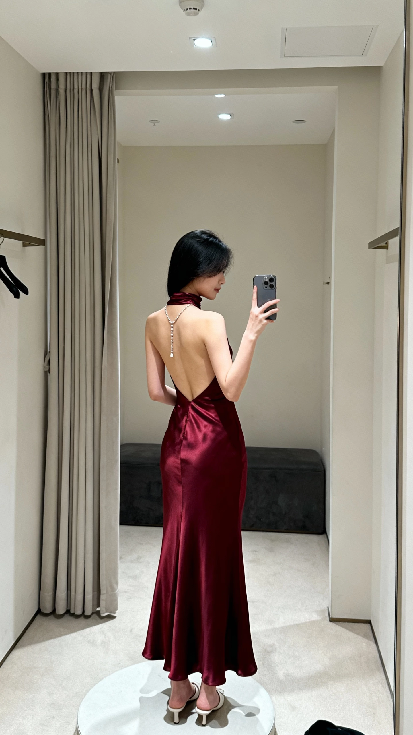iPhone mirror selfie of a woman in a burgundy high-neck satin gown with an open back, drop-back necklace, minimalist heels, face not visible, dressing room mirror, soft overhead lighting, natural iPhone aesthetic.