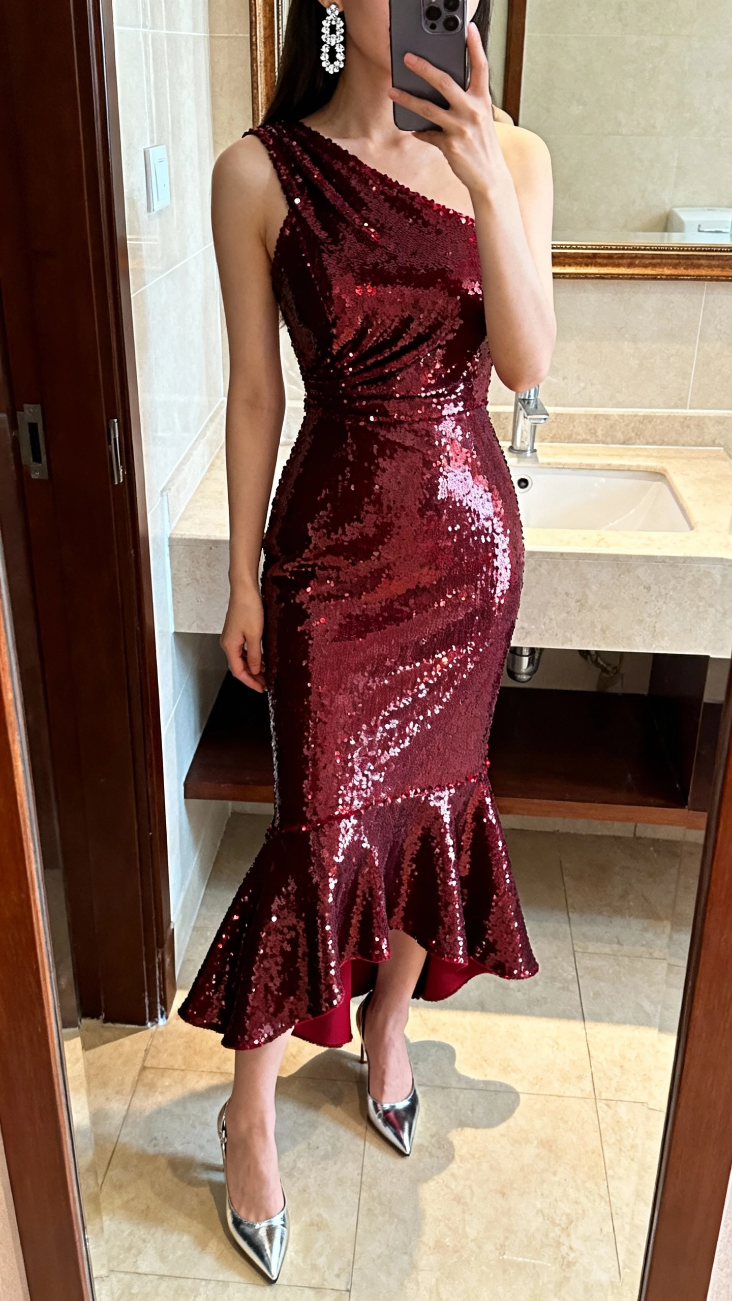 iPhone mirror selfie of a woman in a burgundy sequin one-shoulder gown with a fishtail hem, rhinestone earrings, metallic heels, face not visible, bathroom mirror, warm indoor lighting, natural iPhone photo.