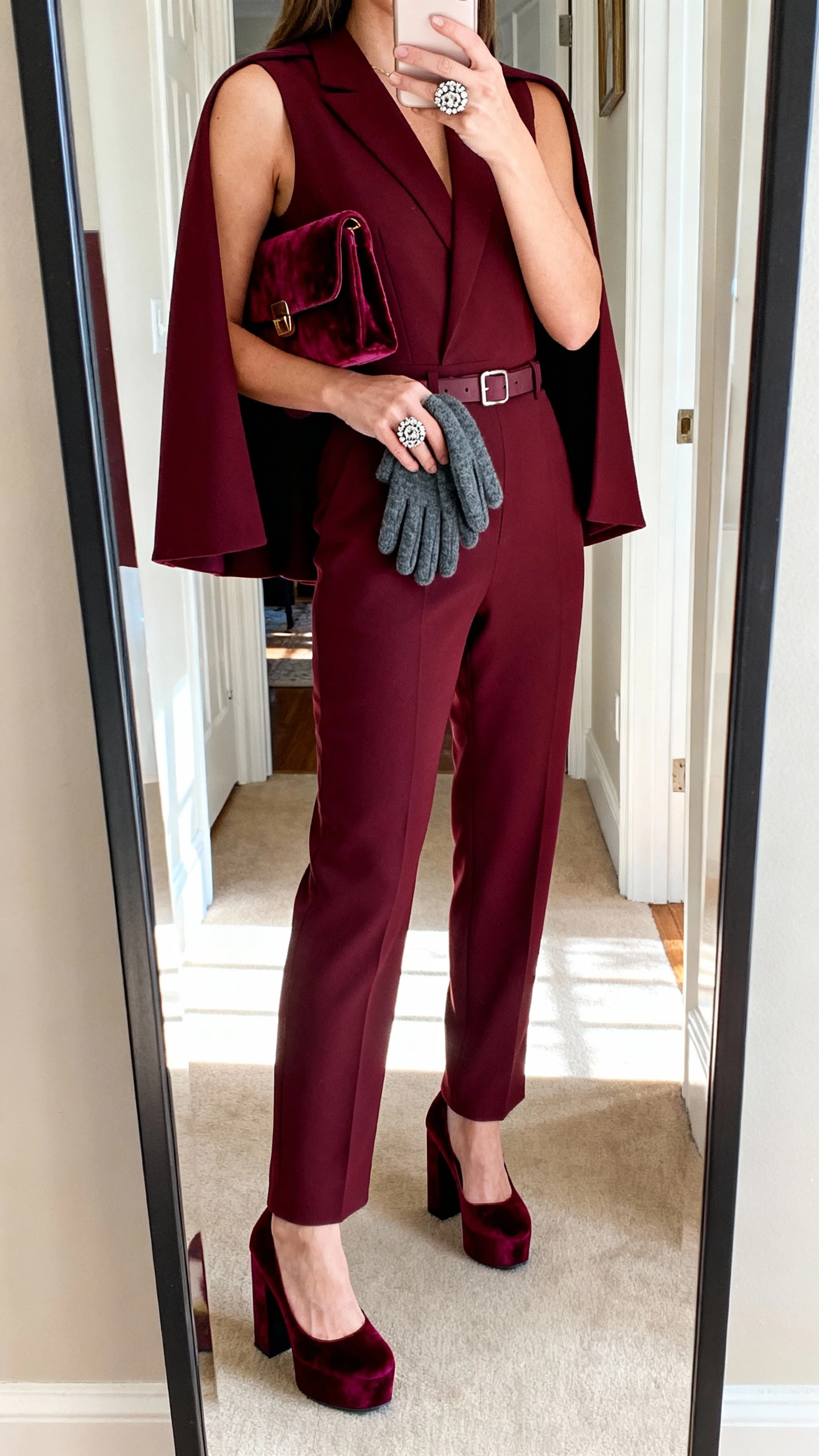 iPhone mirror selfie of a woman in a burgundy sleeveless tailored jumpsuit with matching mini-cape, burgundy velvet platform heels, statement ring, matching velvet clutch, wool gloves tucked into belt; face not visible, entryway mirror, afternoon daylight, casual iPhone photo.
