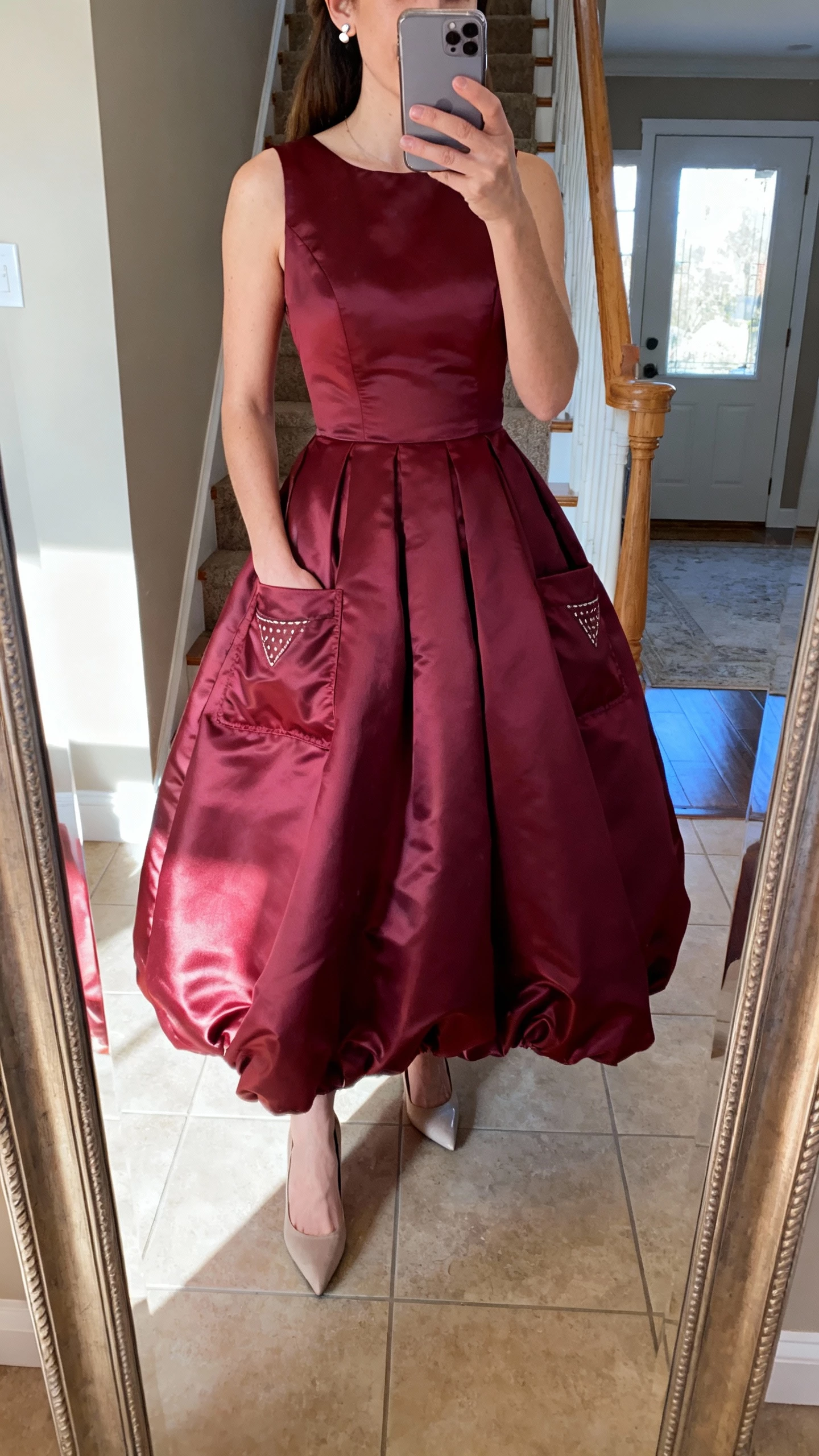 iPhone mirror selfie of a woman in a burgundy taffeta ball gown with pockets, subtle stud earrings, classic pumps, hands in pockets, face not visible, foyer mirror, daylight, casual iPhone aesthetic.