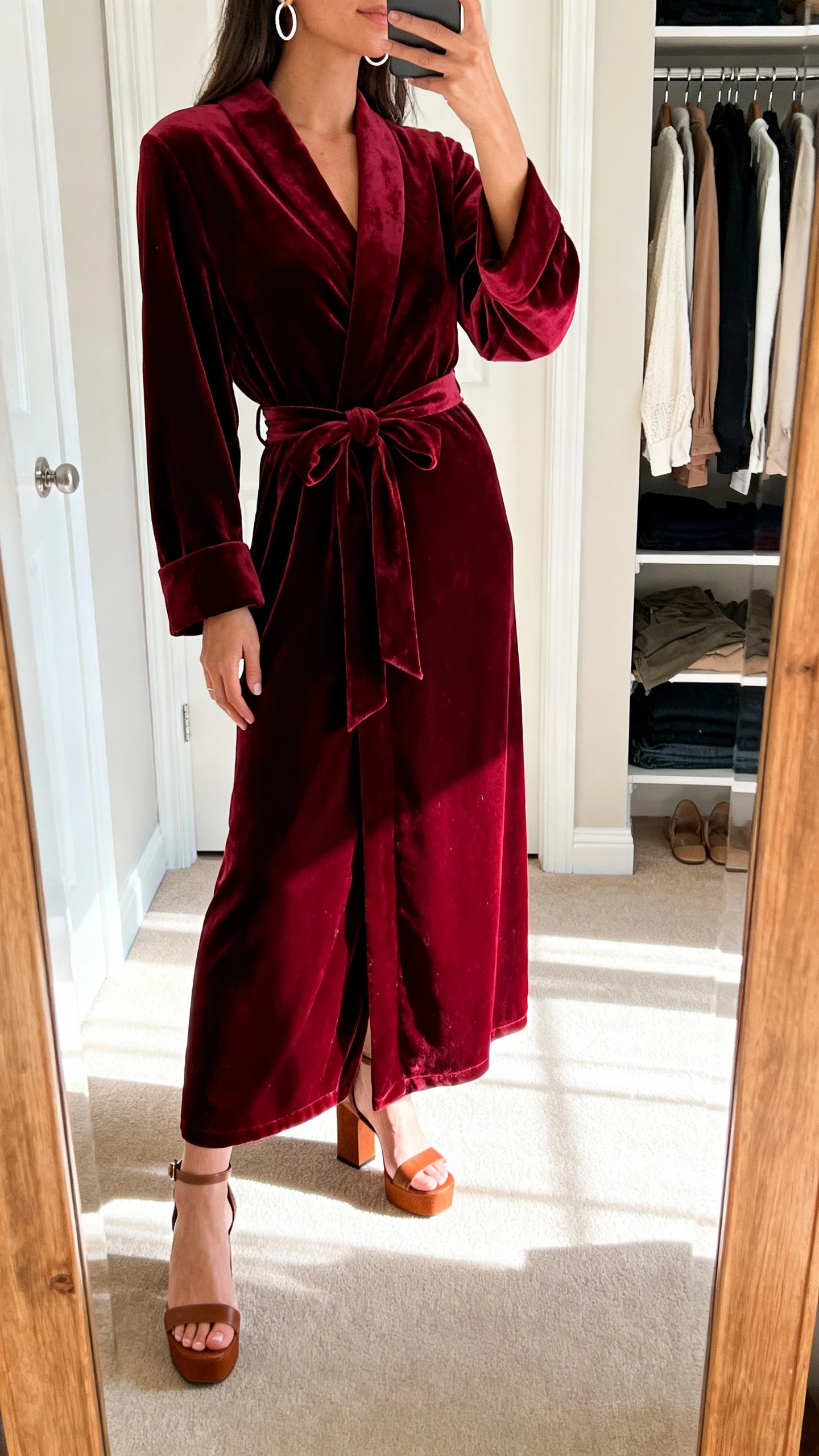 iPhone mirror selfie of a woman in a burgundy velvet wrap gown styled with a waist tie, hoop earrings, block-heel sandals, face not visible, closet mirror, soft afternoon light, natural iPhone aesthetic.