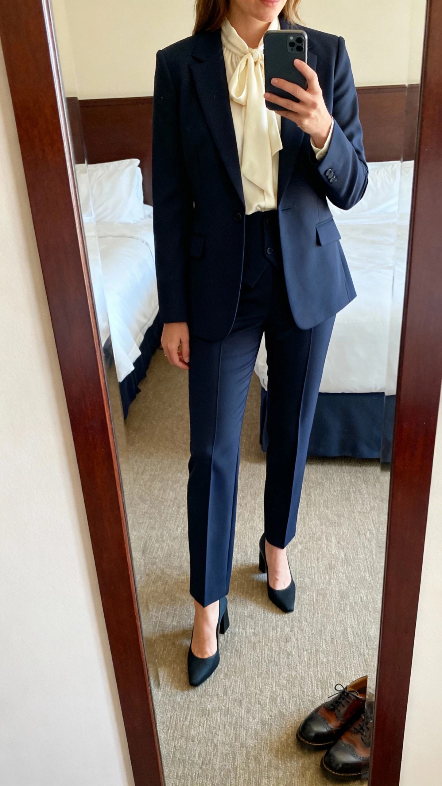 iPhone mirror selfie of a woman in a deep navy three-piece suit: tailored blazer, matching waistcoat (slightly shorter), straight trousers with a crisp crease, cream silk blouse, block-heel pumps, dressy brogues peeking in frame; hotel room mirror, face not visible (cropped at neck), morning natural light, natural iPhone photo quality.