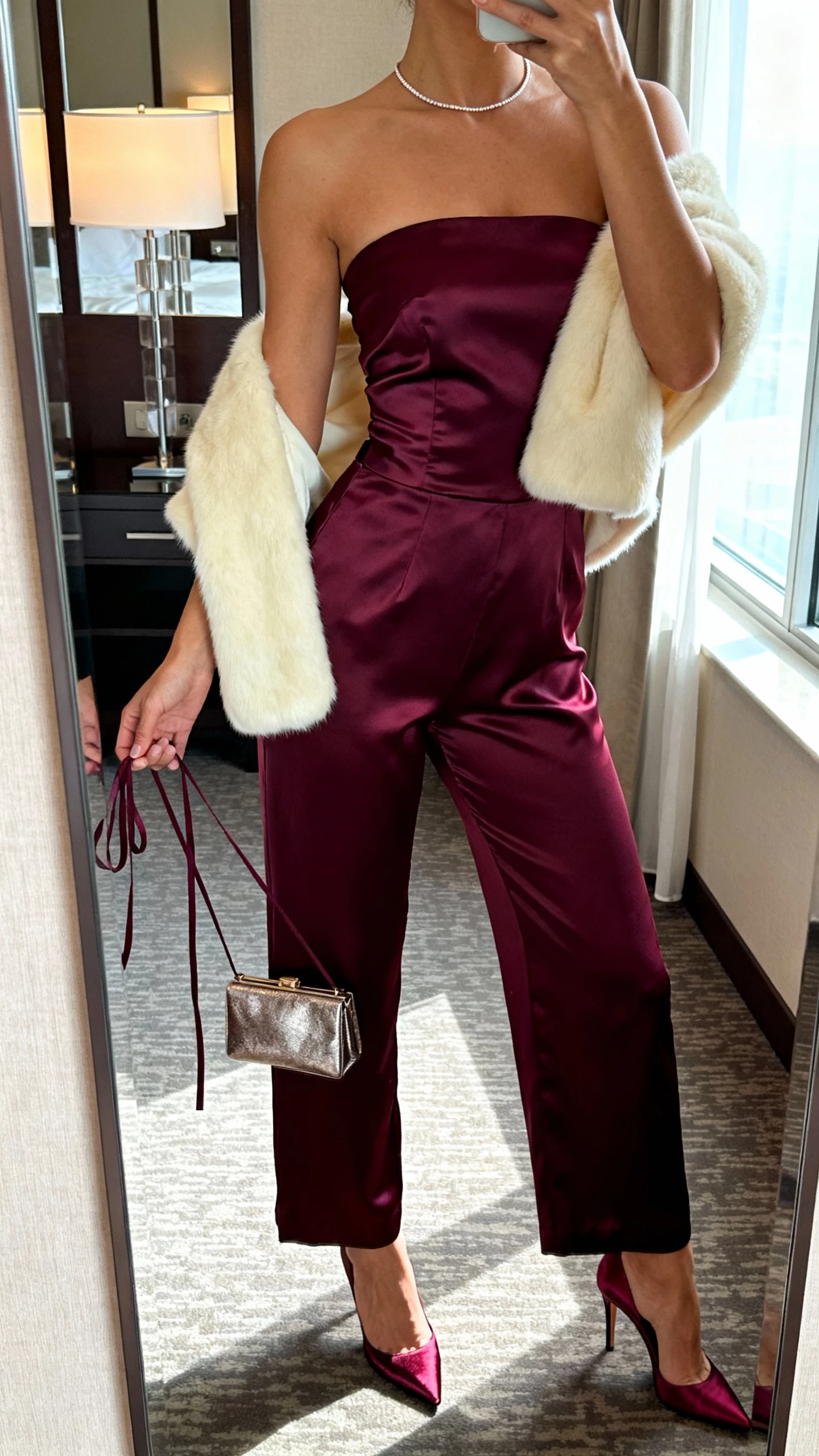iPhone mirror selfie of a woman in a deep plum strapless satin bustier jumpsuit with detachable thin straps held in hand, matching satin stilettos, slim tennis necklace, metallic minaudière, cream short faux-fur shrug over shoulders; face not visible, hotel room mirror, soft lamp and window light, natural iPhone photo.