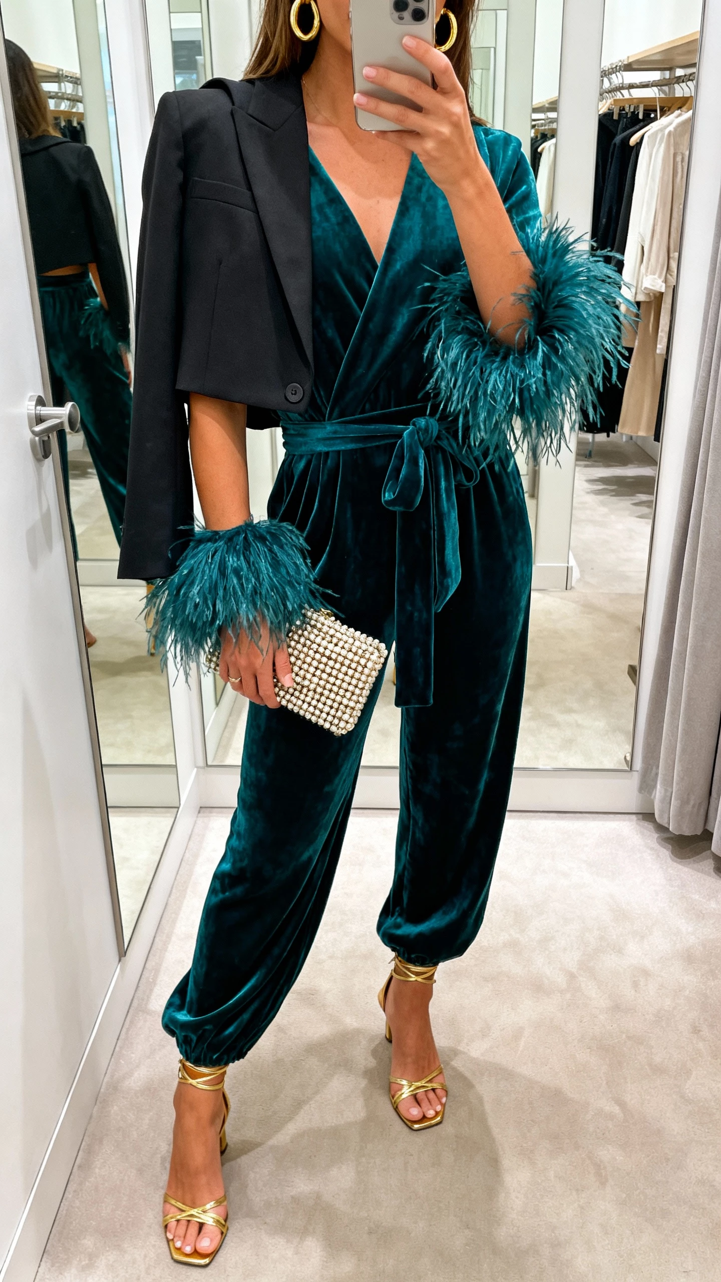 iPhone mirror selfie of a woman in a deep teal velvet wrap jumpsuit with detachable feather cuffs, gold strappy heels, gold hoop earrings, beaded clutch, cropped black tailored blazer over one shoulder; face not visible, boutique fitting room, soft ambient lighting, natural iPhone aesthetic.