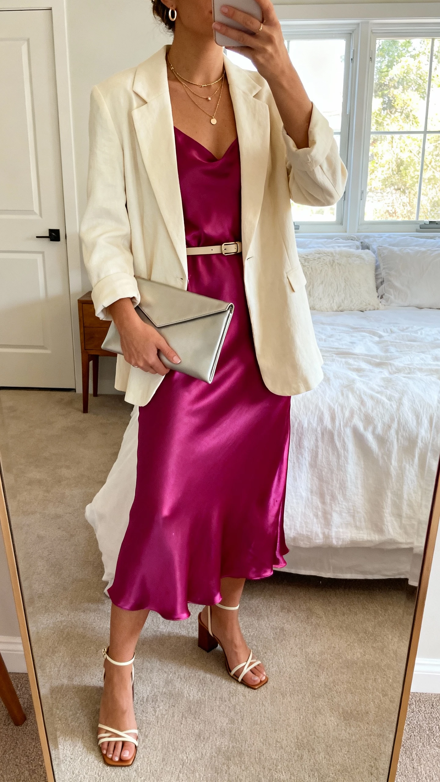 iPhone mirror selfie of a woman in a jewel-tone satin midi slip dress with a cream soft blazer, minimal strappy block-heel sandals, layered necklaces, small hoops, sleek envelope clutch, optional slim waist belt, face not visible, bedroom mirror, natural window light, natural iPhone photo quality.