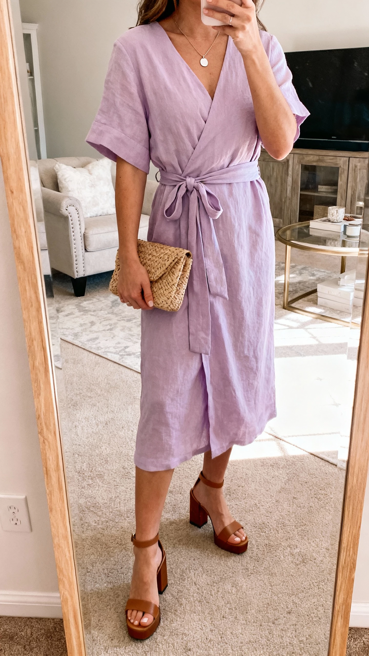 iPhone mirror selfie of a woman in a lavender wrap dress with tie waist, block-heel sandals, a simple pendant necklace, and a small woven clutch, face not visible, full-length mirror in living room, soft afternoon light, natural iPhone photo quality.