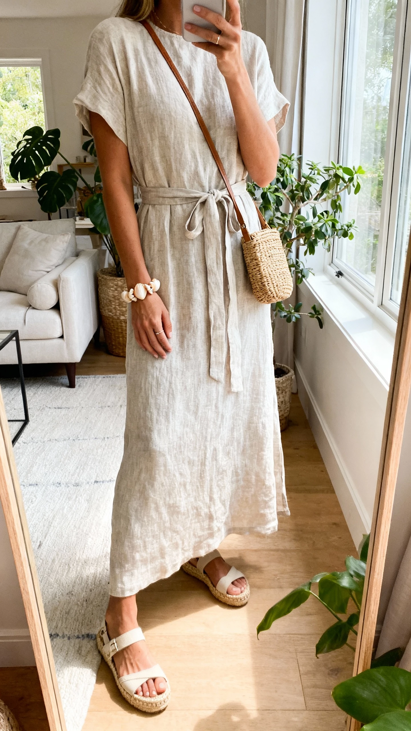 iPhone mirror selfie of a woman in a linen-blend maxi dress with a soft waist tie, espadrille wedges, shell bracelet, and a rattan crossbody, face not visible, airy living room mirror with plants, morning window light, natural iPhone aesthetic