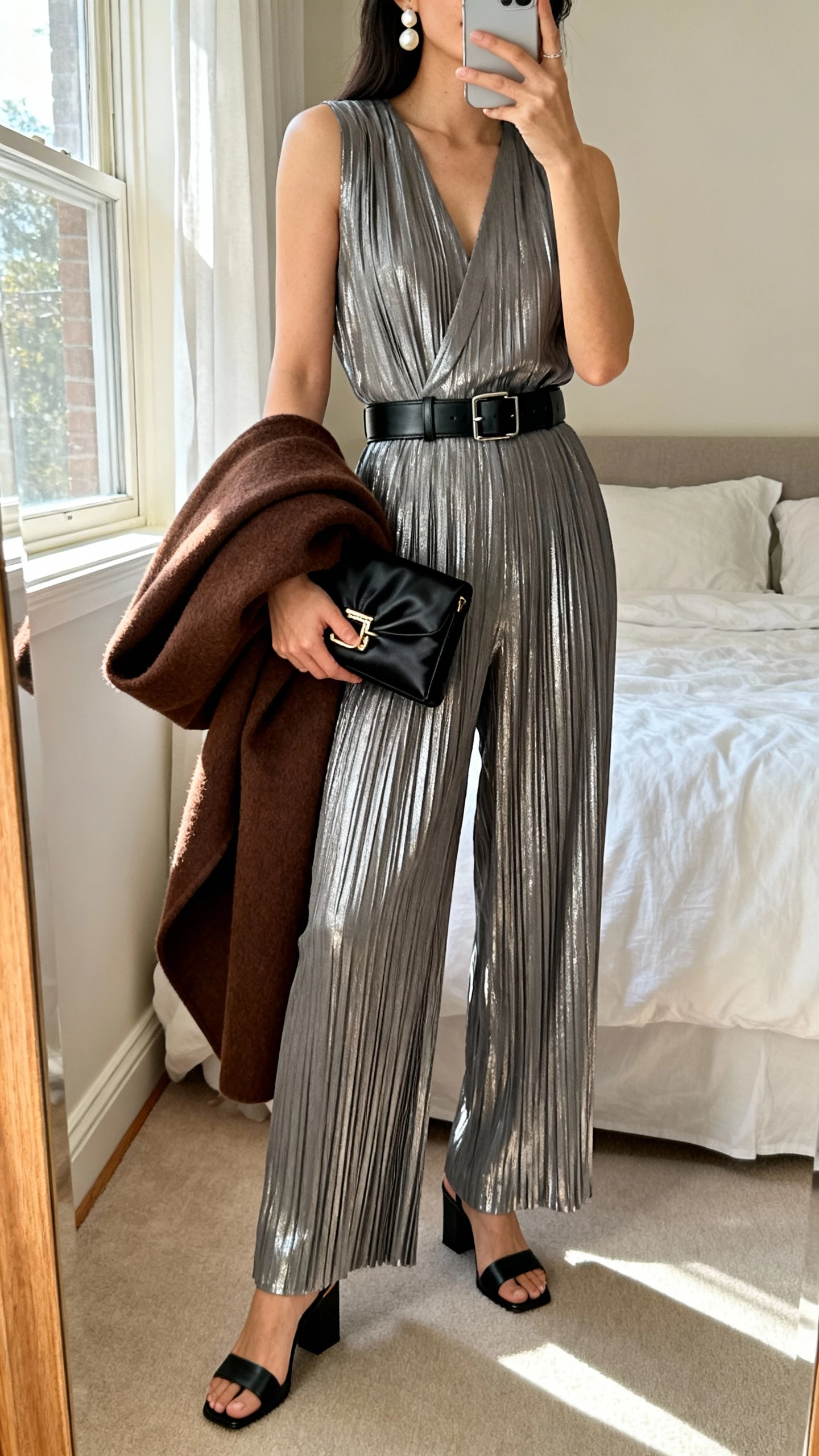 iPhone mirror selfie of a woman in a pewter metallic pleated wide-leg jumpsuit, matte black leather belt, black block-heel sandals, drop pearl earrings, black satin frame-clasp clutch, chocolate brown wool wrap coat folded over arm; face not visible, bedroom full-length mirror, afternoon window light, casual iPhone photo.