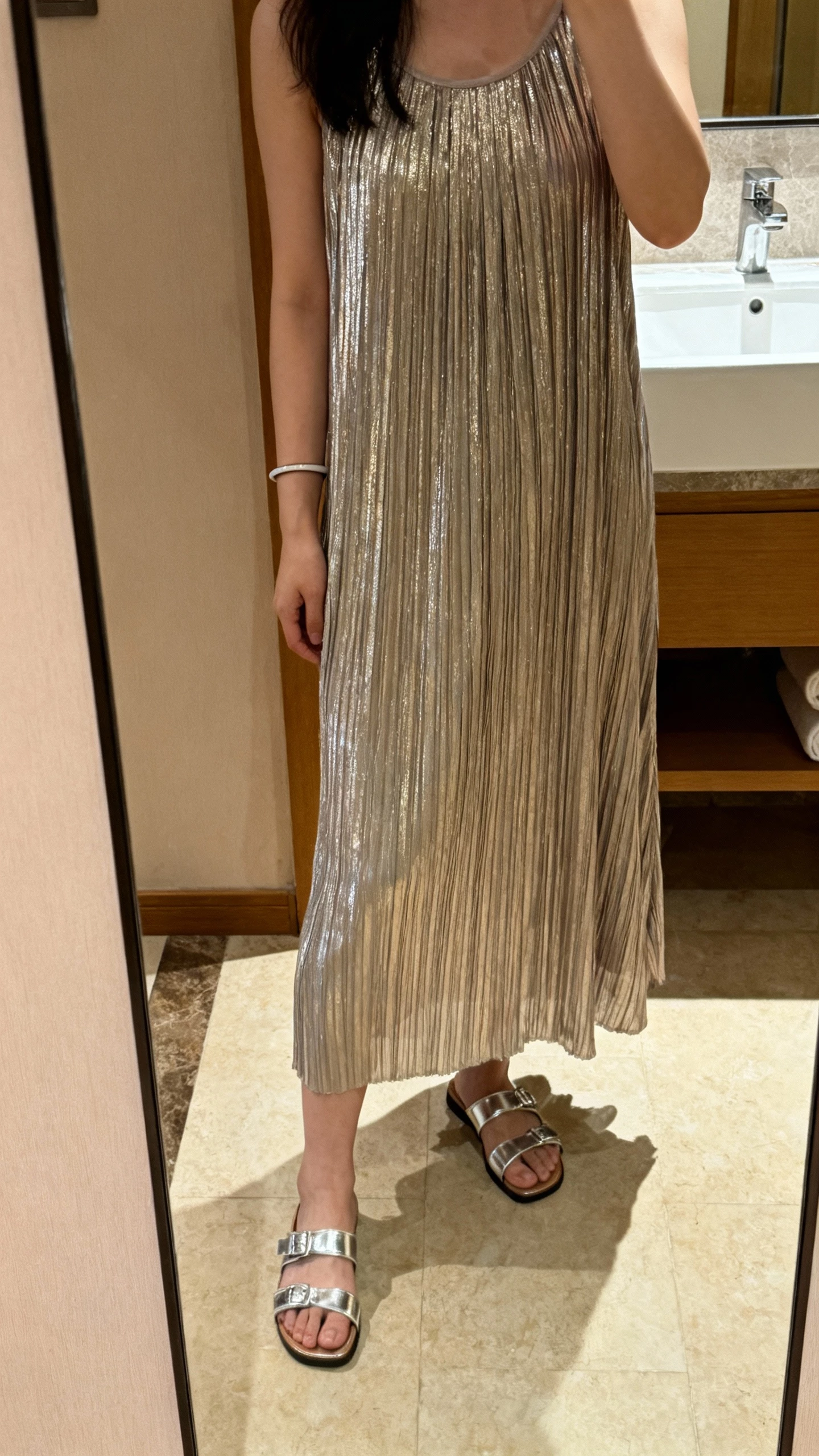 iPhone mirror selfie of a woman in a pleated shimmery midi dress with metallic sandals and a slim bracelet, face not visible, cropped at shoulders, hotel bathroom mirror, warm indoor lighting, natural iPhone look