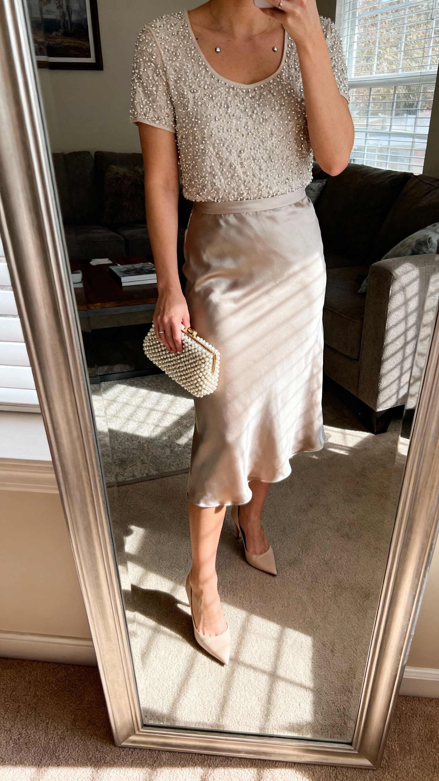 iPhone mirror selfie of a woman in a silk midi skirt paired with a subtly beaded top, kitten heels, beaded clutch, understated studs, face not visible, cropped at neck in a living room mirror with afternoon window light, natural iPhone photo quality