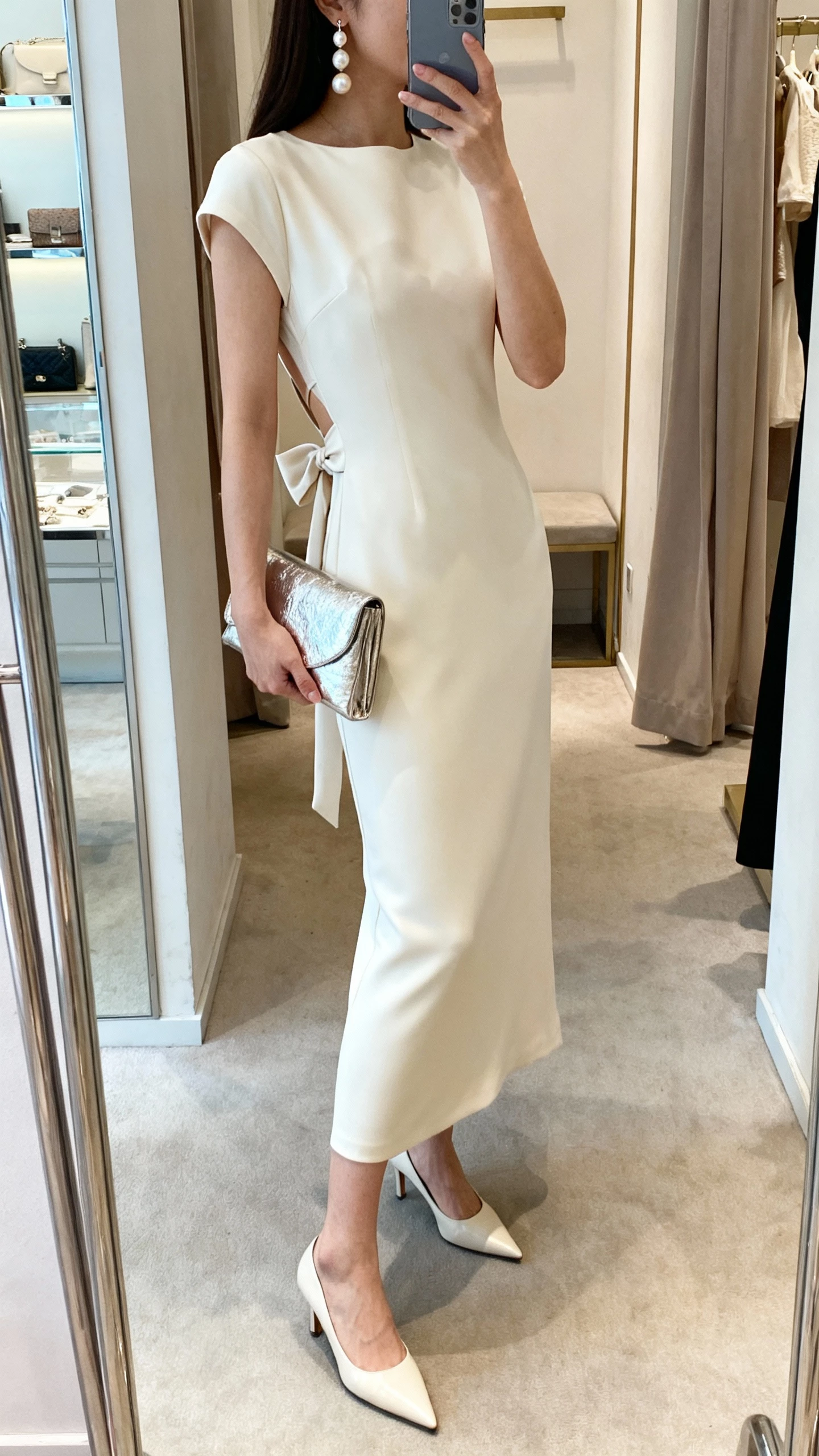 iPhone mirror selfie of a woman in a sleek column dress with a bow-back detail, pointed-toe heels, pearl drop earrings, and a metallic clutch, face not visible, boutique fitting room mirror, soft indoor lighting, natural iPhone photo quality.