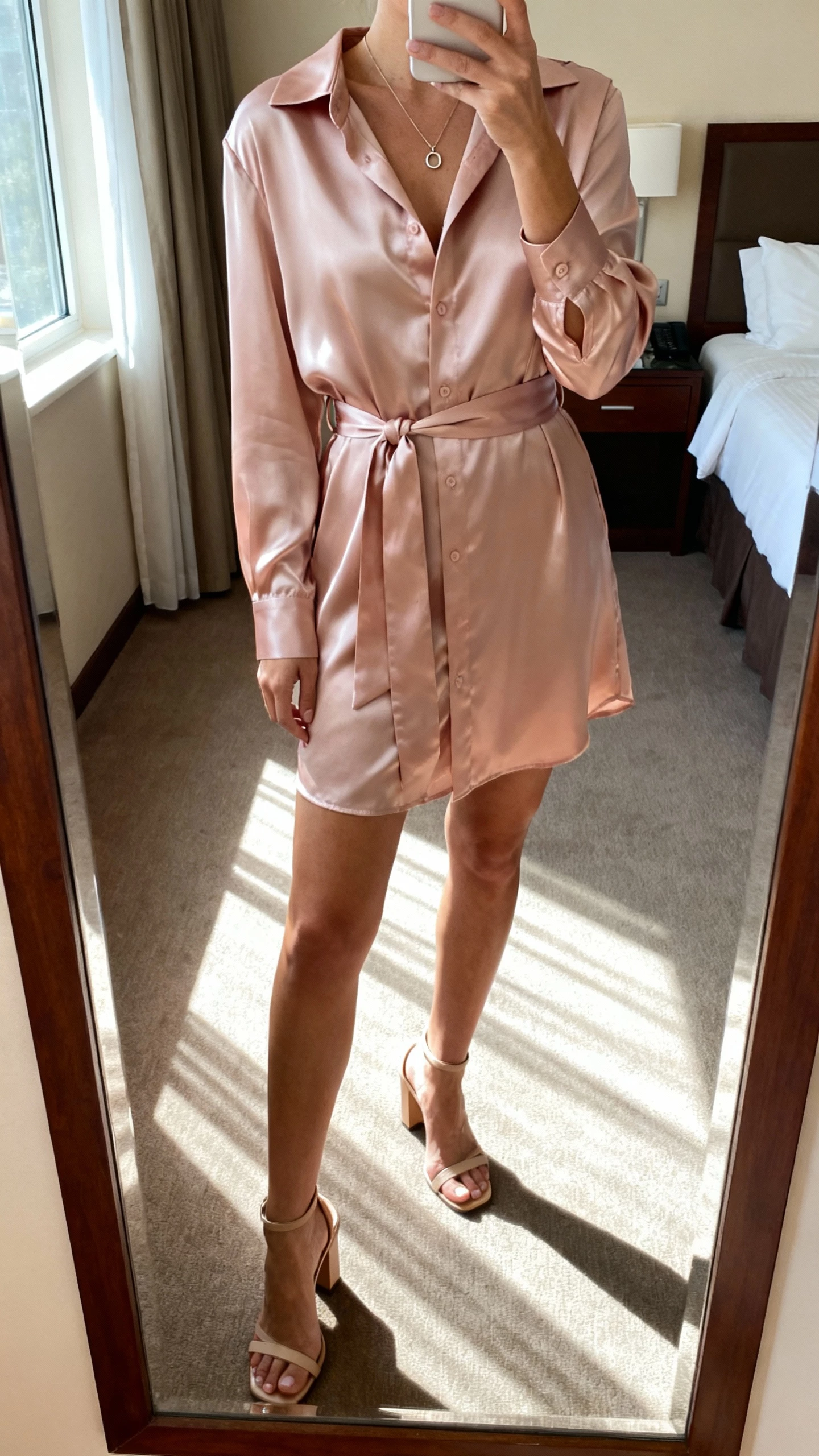 iPhone mirror selfie of a woman in a soft blush satin shirt dress with a waist tie, simple pendant, heeled sandals, face not visible, cropped at shoulders, hotel room mirror, morning window light, casual iPhone aesthetic
