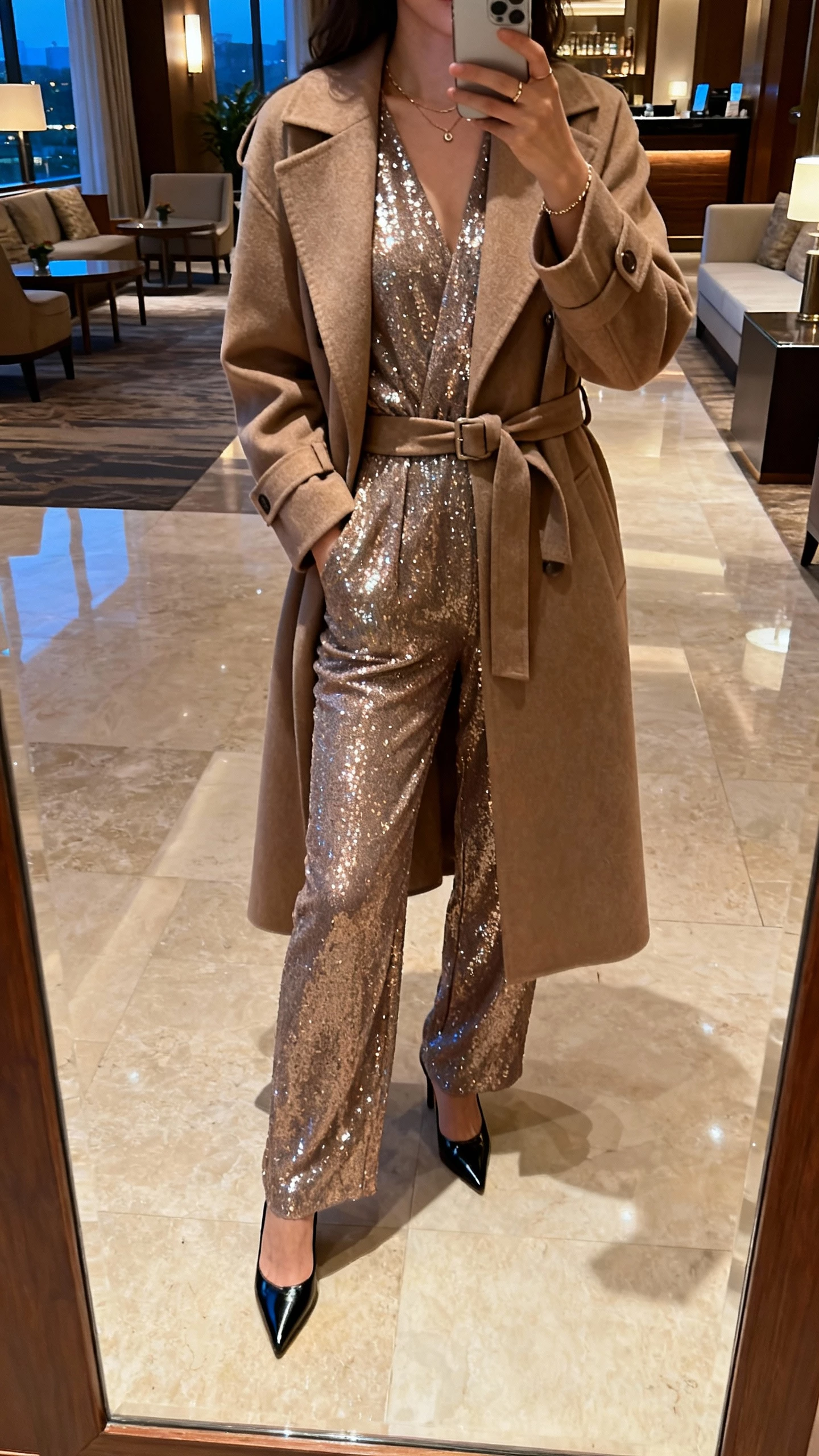 iPhone mirror selfie of a woman in a sparkly jumpsuit under a belted long coat, pointed heels, minimal jewelry, face not visible, hotel lobby mirror, ambient evening light, natural iPhone look.