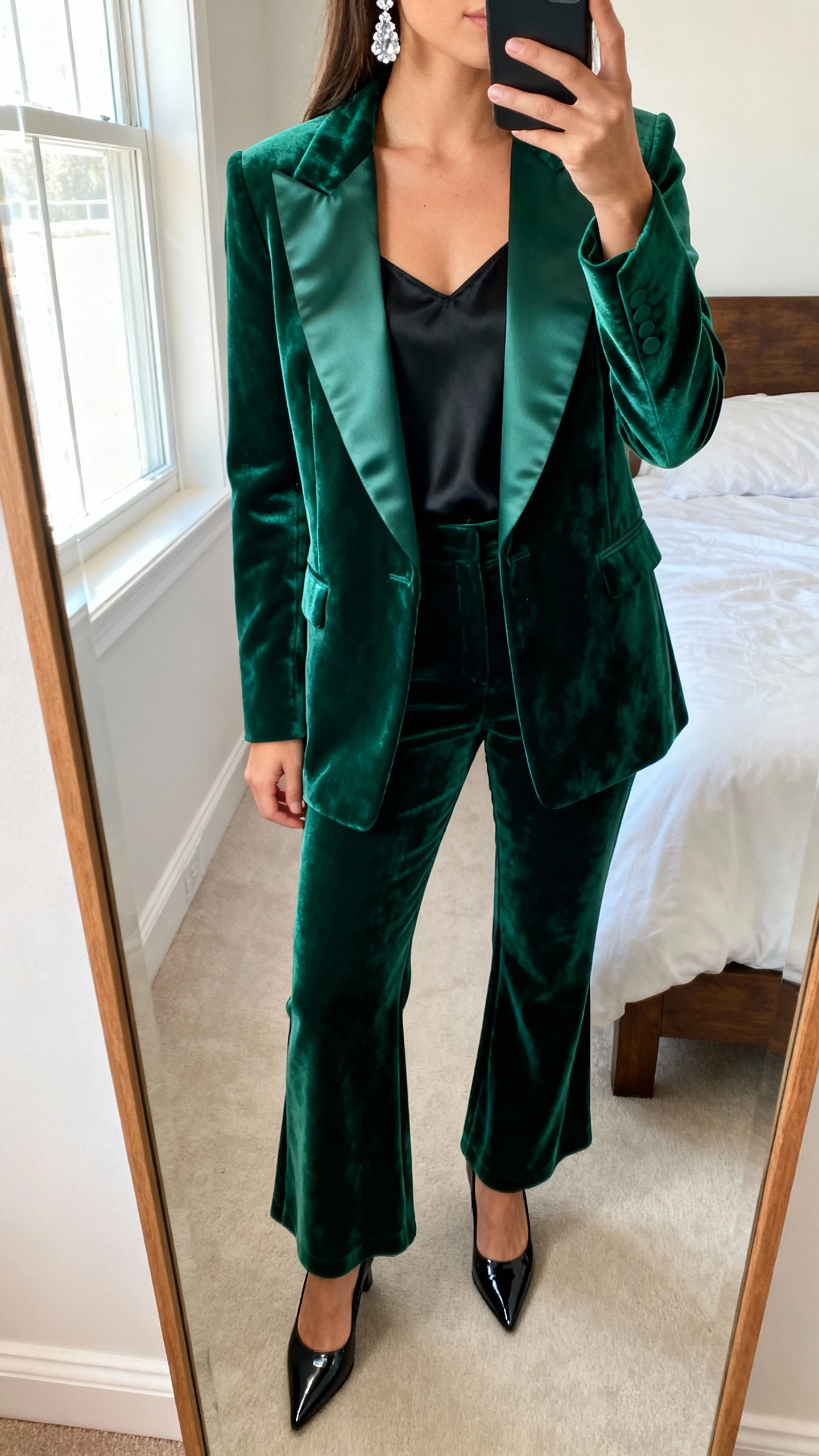 iPhone mirror selfie of a woman in an emerald velvet tuxedo blazer with satin lapels, matching velvet tailored trousers (slight flare), black silk camisole, black patent pointed-toe heels, crystal drop earrings; bedroom mirror, face not visible (cropped at neck), natural window light, natural iPhone photo quality.