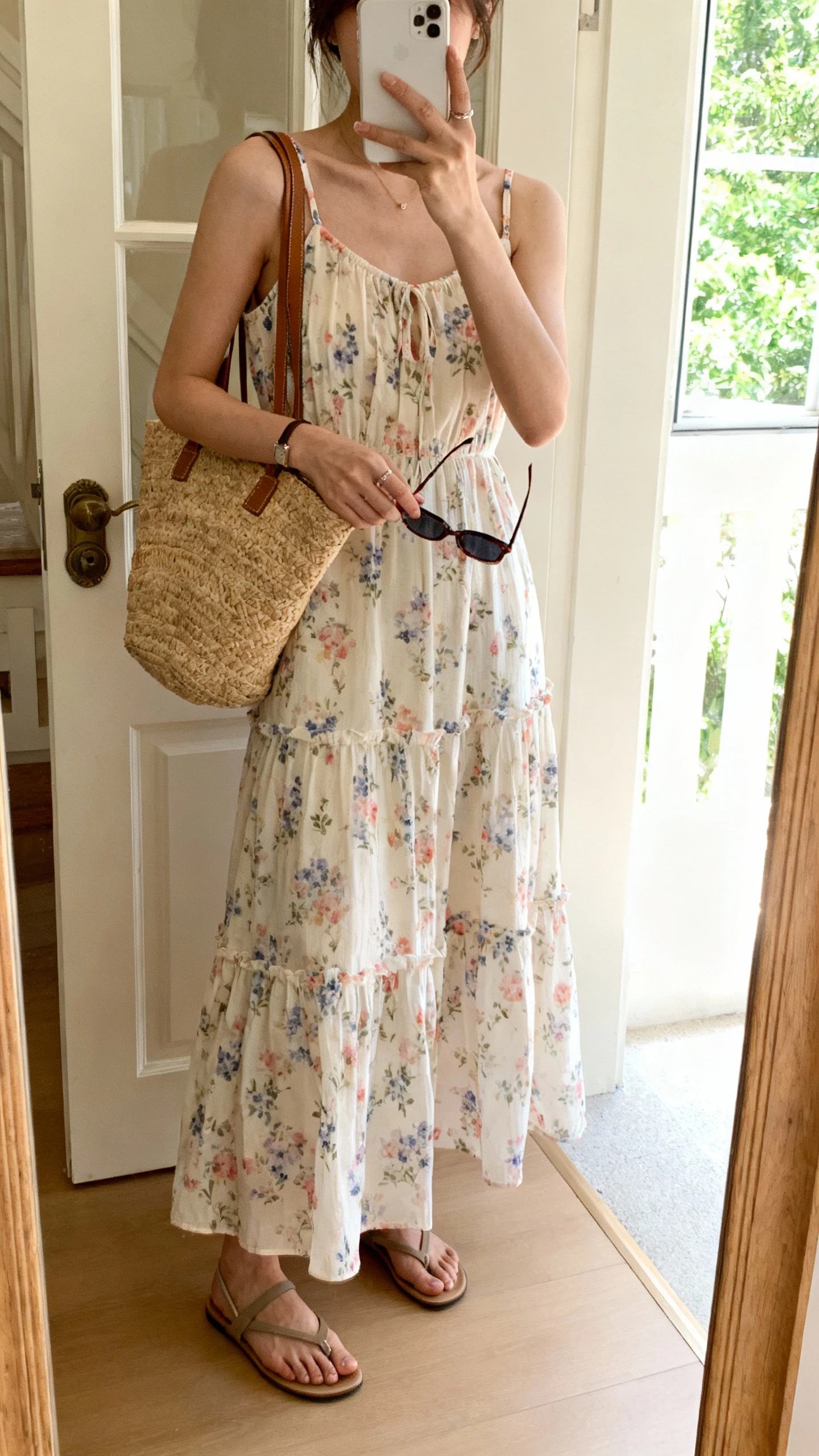 iPhone mirror selfie of a woman wearing a breezy tiered floral sundress with straps, flat sandals, straw tote, and sunglasses in hand, face not visible, entryway mirror by the door, bright daylight from window, casual natural iPhone photo.