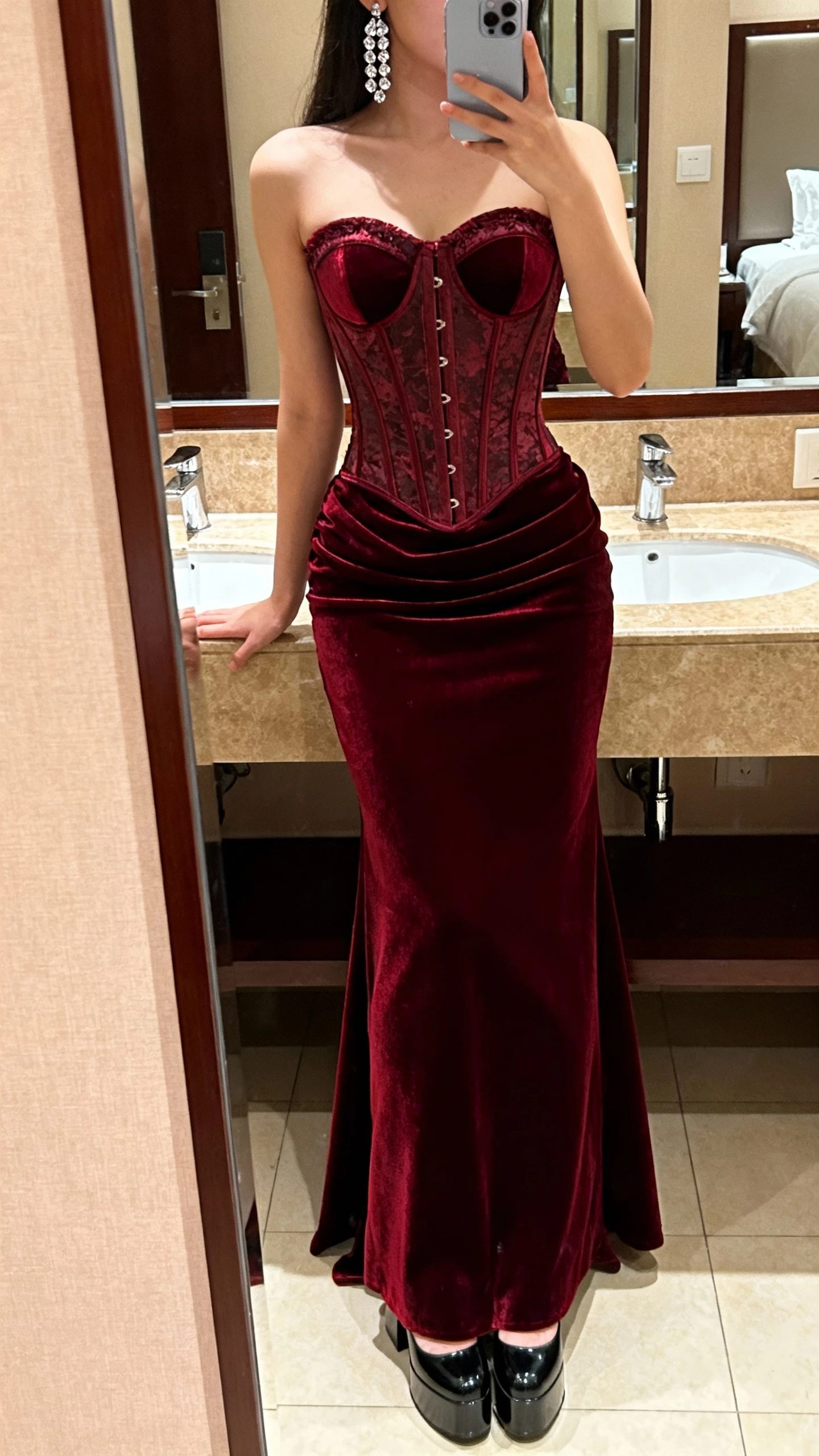 iPhone mirror selfie of a woman wearing a burgundy velvet corset gown with bustier detail, crystal earrings, platform heels, face not visible, hotel bathroom mirror, warm indoor lighting, casual iPhone quality.