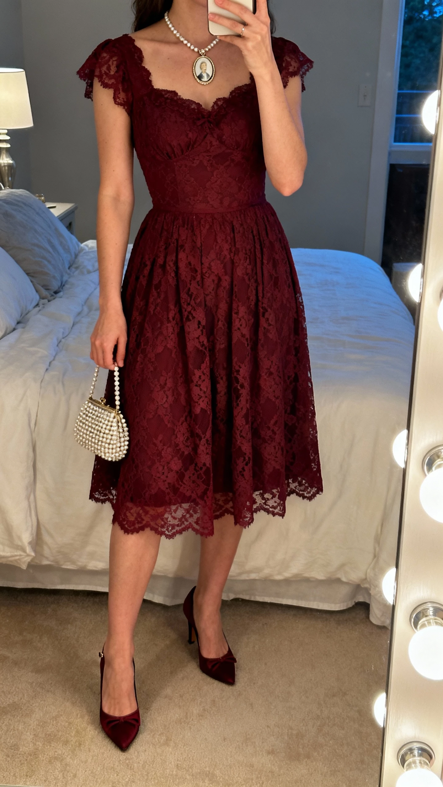 iPhone mirror selfie of a woman wearing a burgundy lace tea-length dress, vintage-inspired kitten heels, cameo necklace, and a small beaded evening bag; face not visible, bedroom mirror with vanity lights, soft evening lamp light, natural iPhone quality.