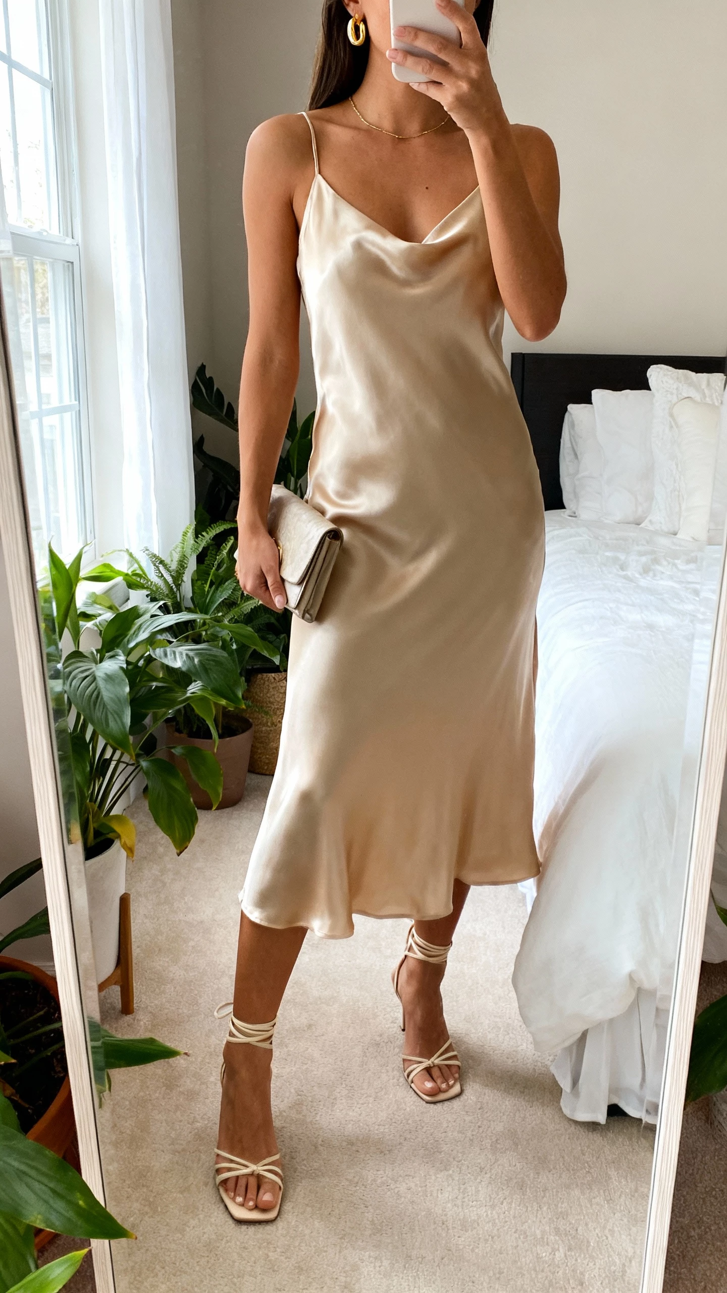 iPhone mirror selfie of a woman wearing a champagne-colored satin slip dress with strappy heels, delicate gold earrings, and a small clutch, face not visible, cropped at neck, bedroom mirror with plants, soft window light, natural iPhone photo quality