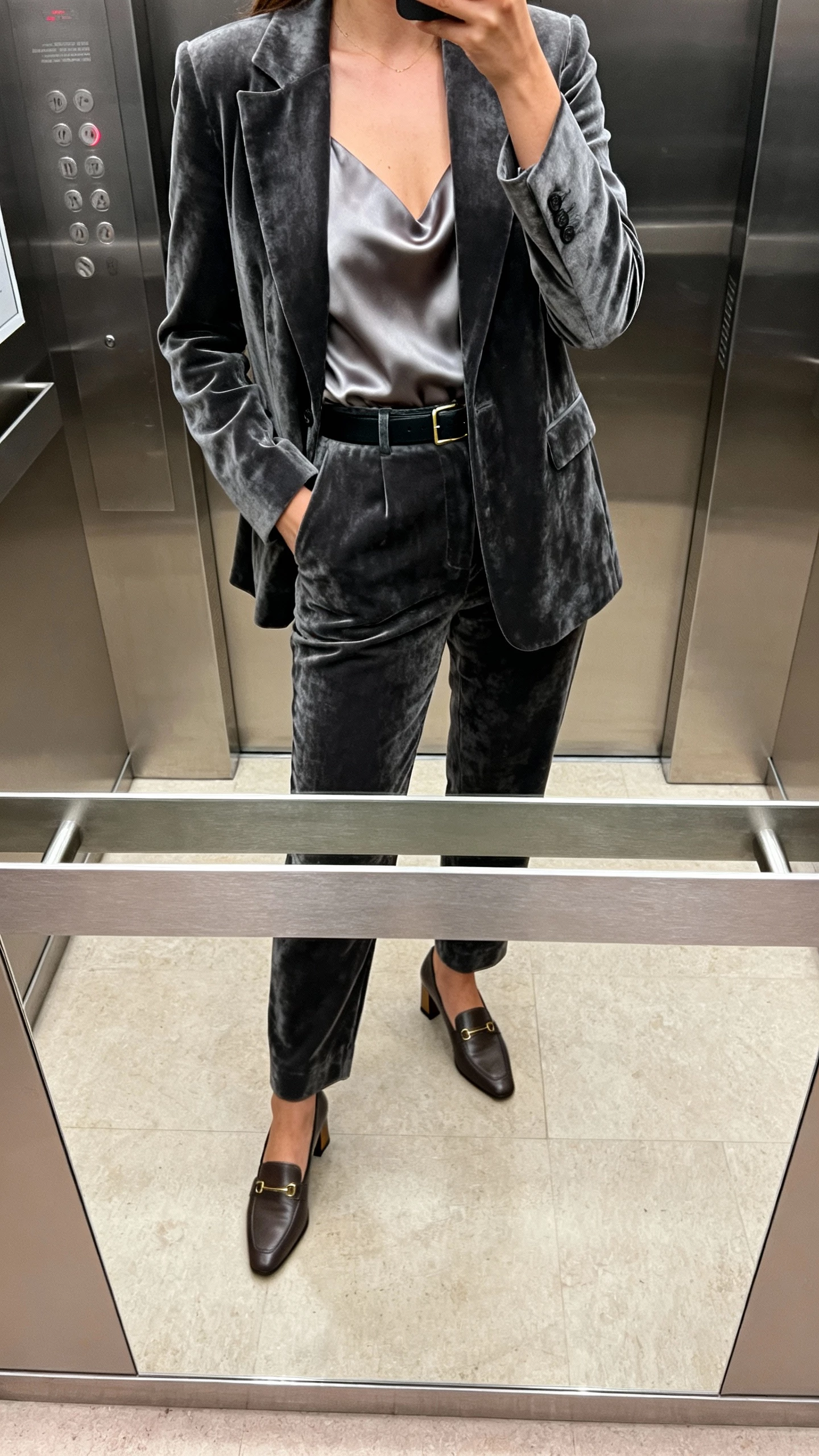 iPhone mirror selfie of a woman wearing a charcoal velvet suit with a silk shell, slim belt, heeled loafers, face not visible, cropped at neck, elevator mirror, ambient indoor lighting, natural iPhone photo quality