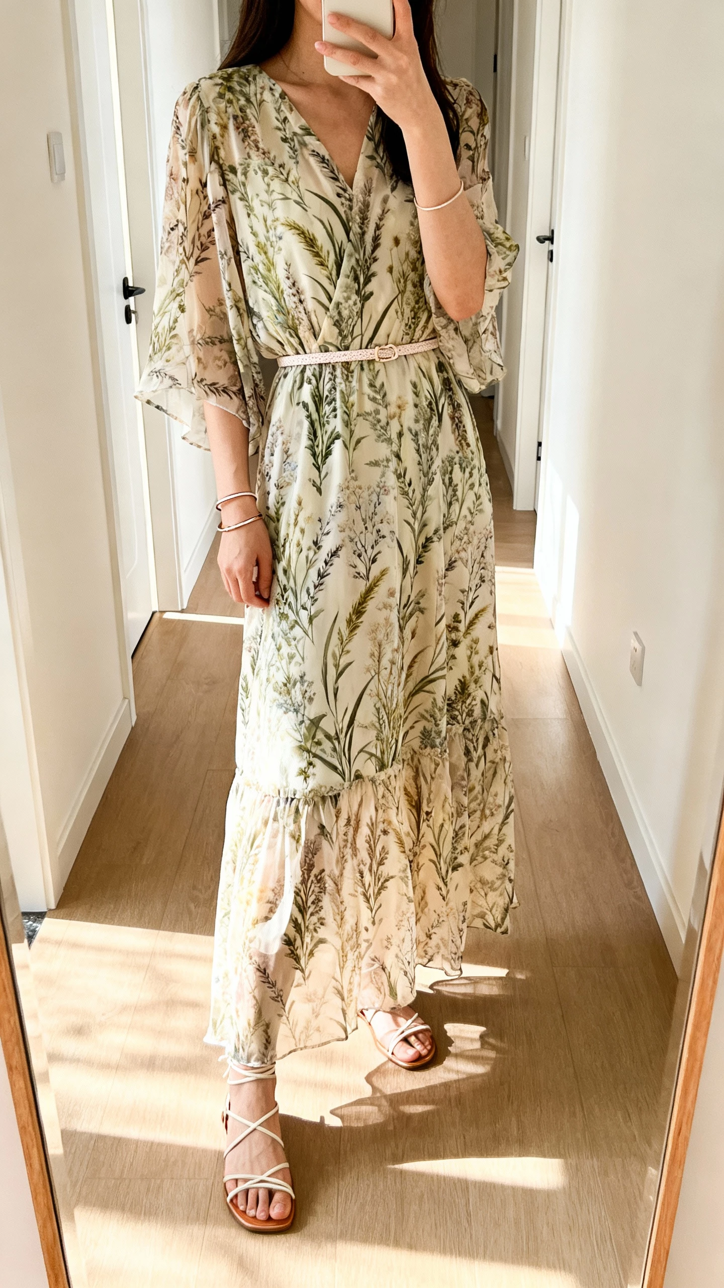 iPhone mirror selfie of a woman wearing a chiffon maxi with meadow print and a delicate belt, thin bangles, strappy sandals, face not visible, taken in a softly lit hallway with natural afternoon light, iPhone photo quality