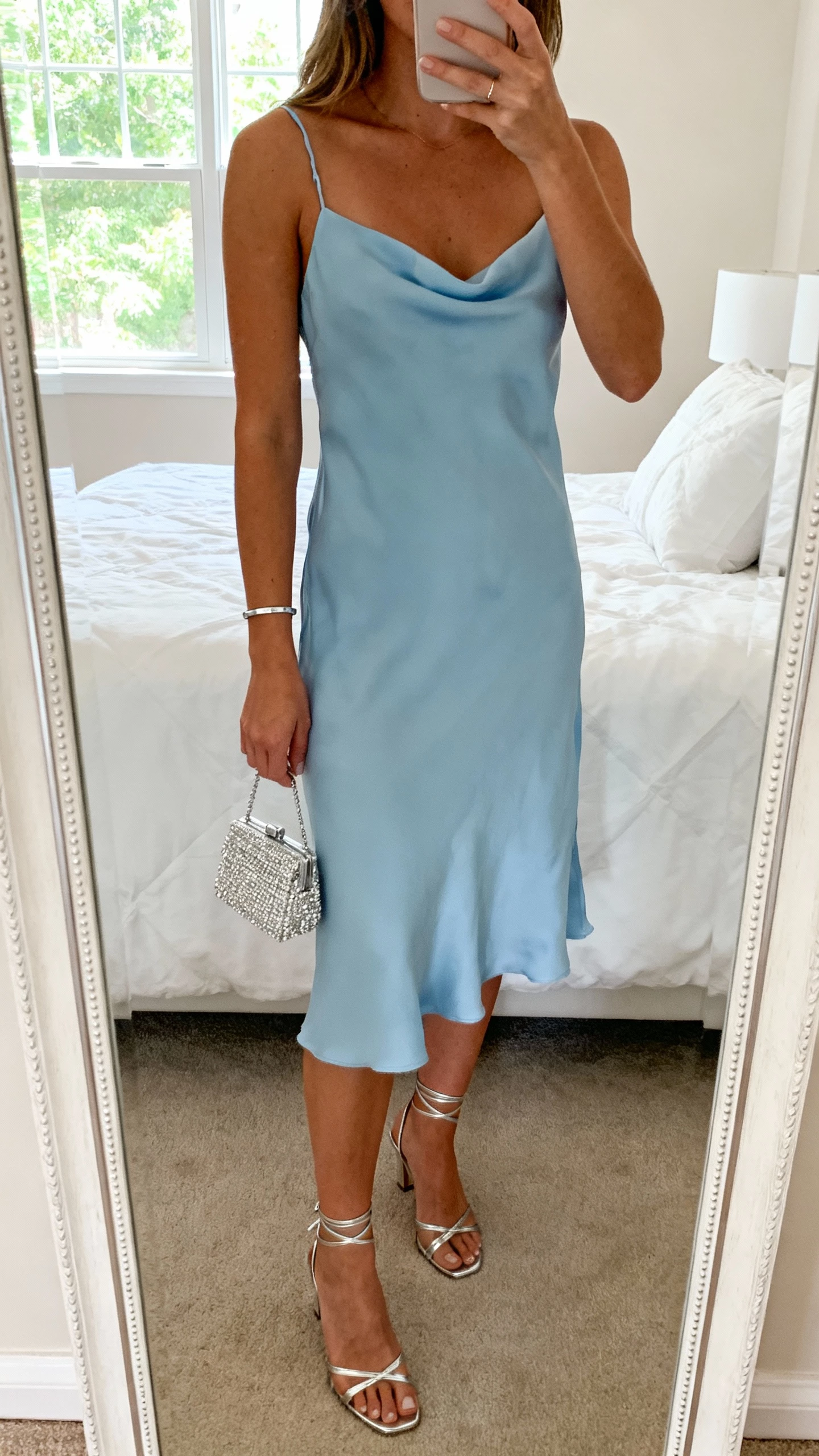 iPhone mirror selfie of a woman wearing a cloud-blue slip dress with delicate spaghetti straps, strappy silver heels, a slim silver bracelet, and a tiny crystal clutch, face not visible, cropped at neck, bedroom mirror with morning window light, natural iPhone photo quality.