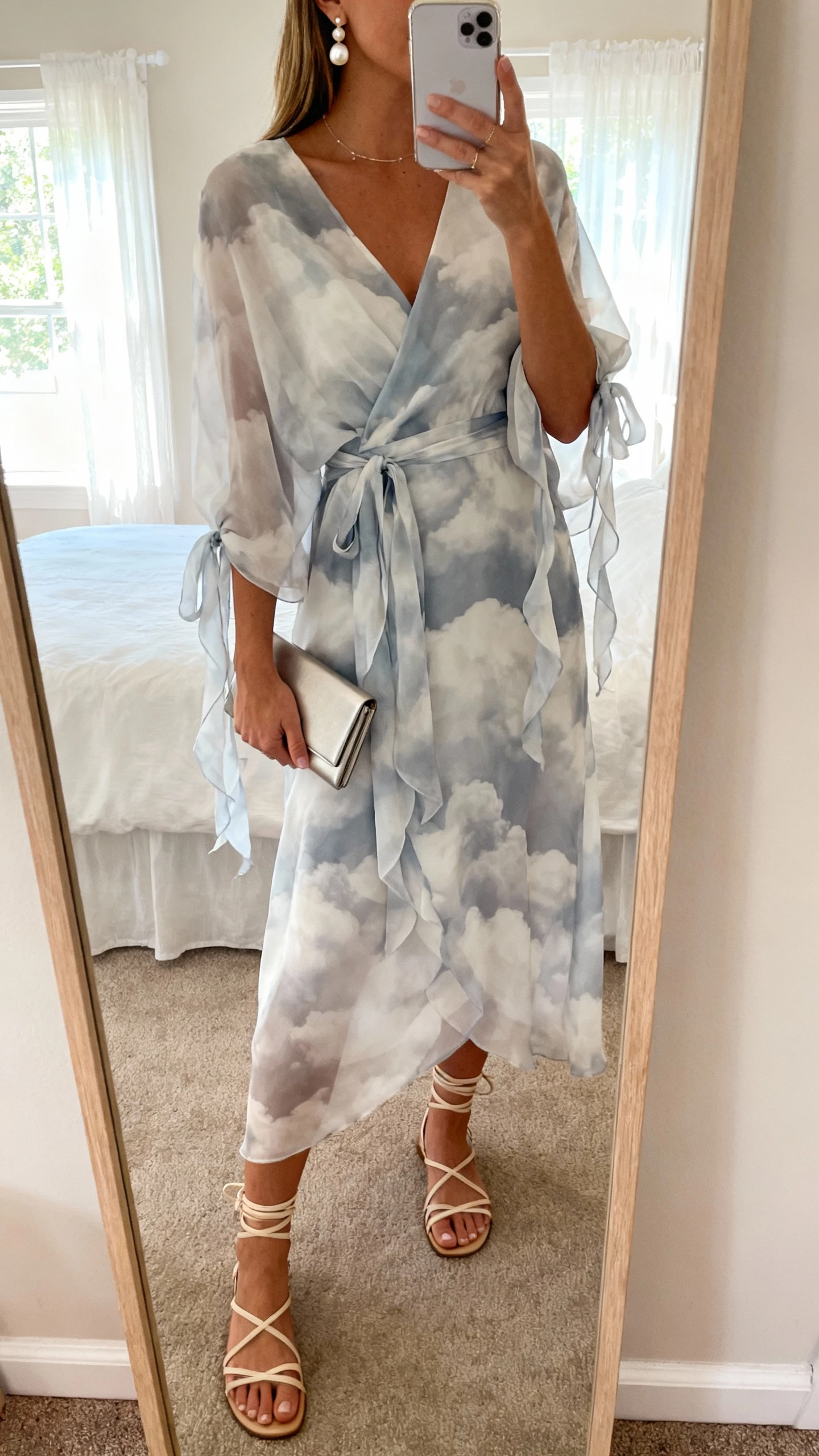 iPhone mirror selfie of a woman wearing a cloud-chiffon wrap dress with floaty ties, strappy sandals, delicate pearl earrings, and a slim clutch, face not visible, cropped at neck, bedroom mirror with soft window light, natural iPhone photo quality