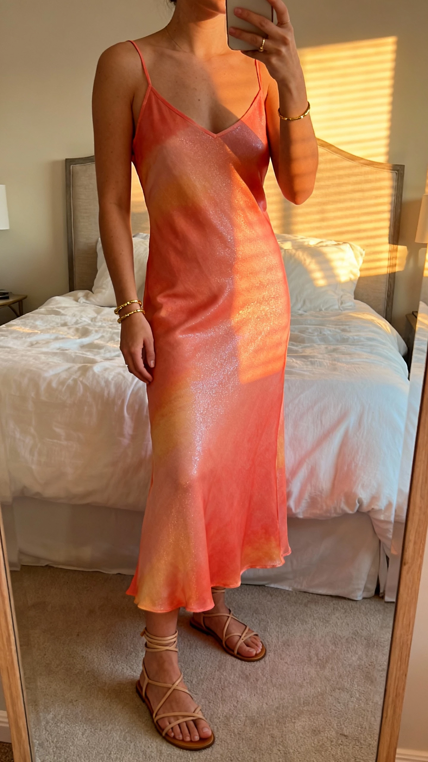 iPhone mirror selfie of a woman wearing a coral sunset slip dress with a subtle shimmer, strappy sandals, delicate gold bracelets, face not visible, cropped at neck, bedroom mirror with golden-hour window light, natural iPhone photo quality.