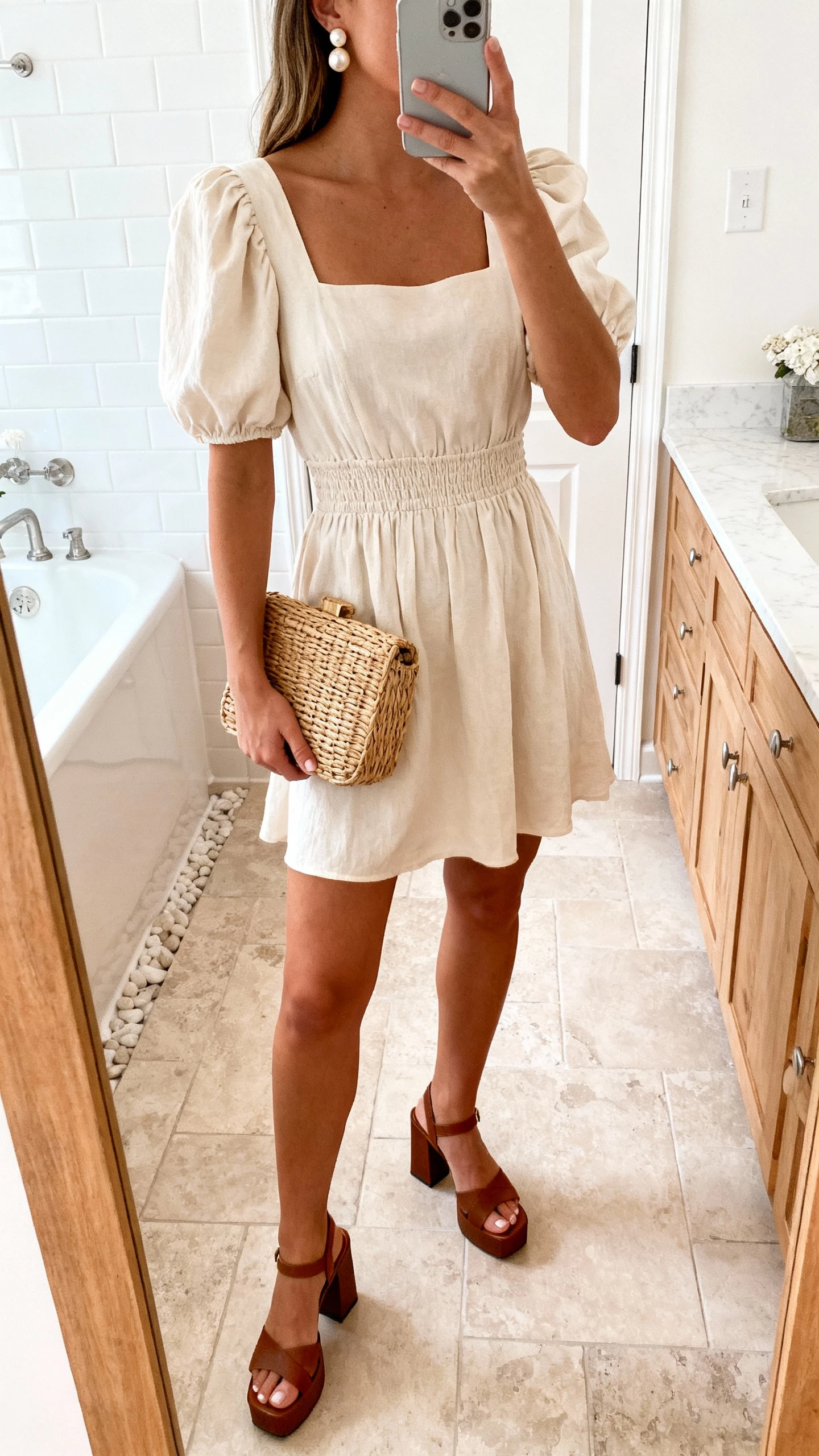 iPhone mirror selfie of a woman wearing a cottagecore puff-sleeve mini dress with a square neckline and smocked waist, block-heel sandals, pearl studs, and a wicker clutch, face not visible, bathroom mirror, bright indoor lighting, natural iPhone photo.