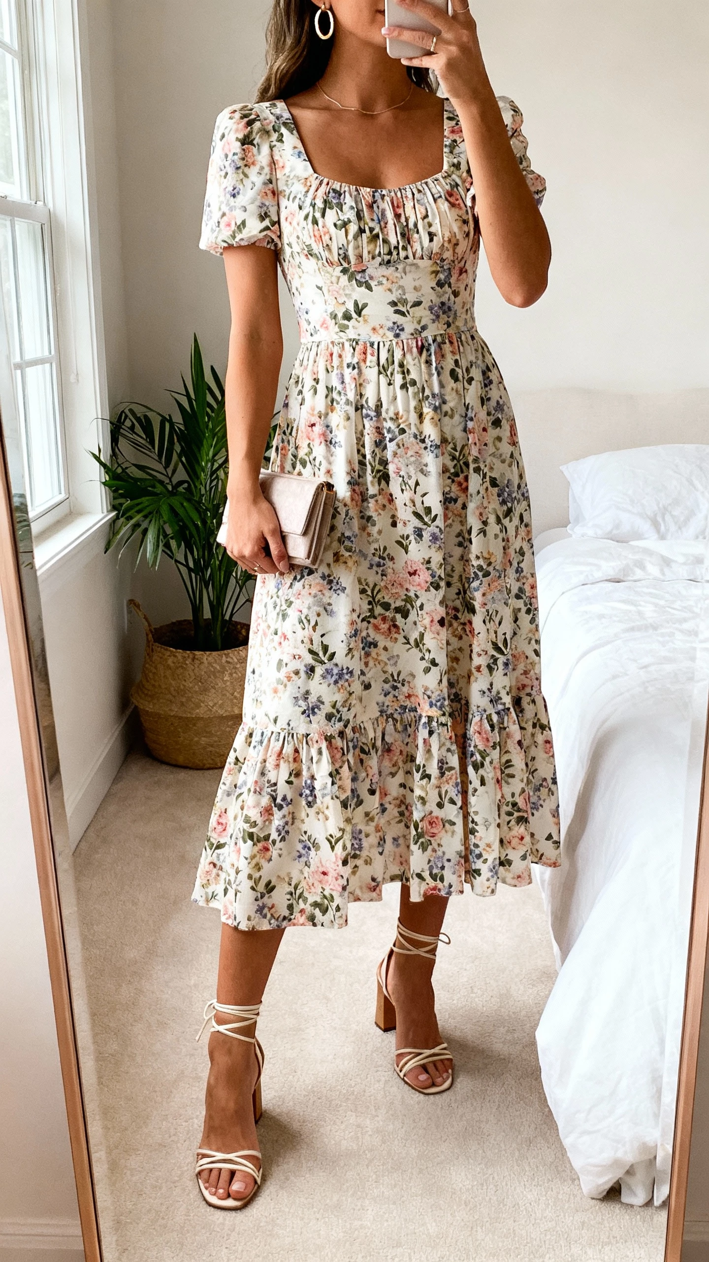 iPhone mirror selfie of a woman wearing a floral garden-party midi dress with a fitted bodice and flowy skirt, strappy heeled sandals, delicate hoop earrings, and a small clutch, face not visible, cropped at neck, bedroom mirror with potted plant, soft window light, natural iPhone photo quality.