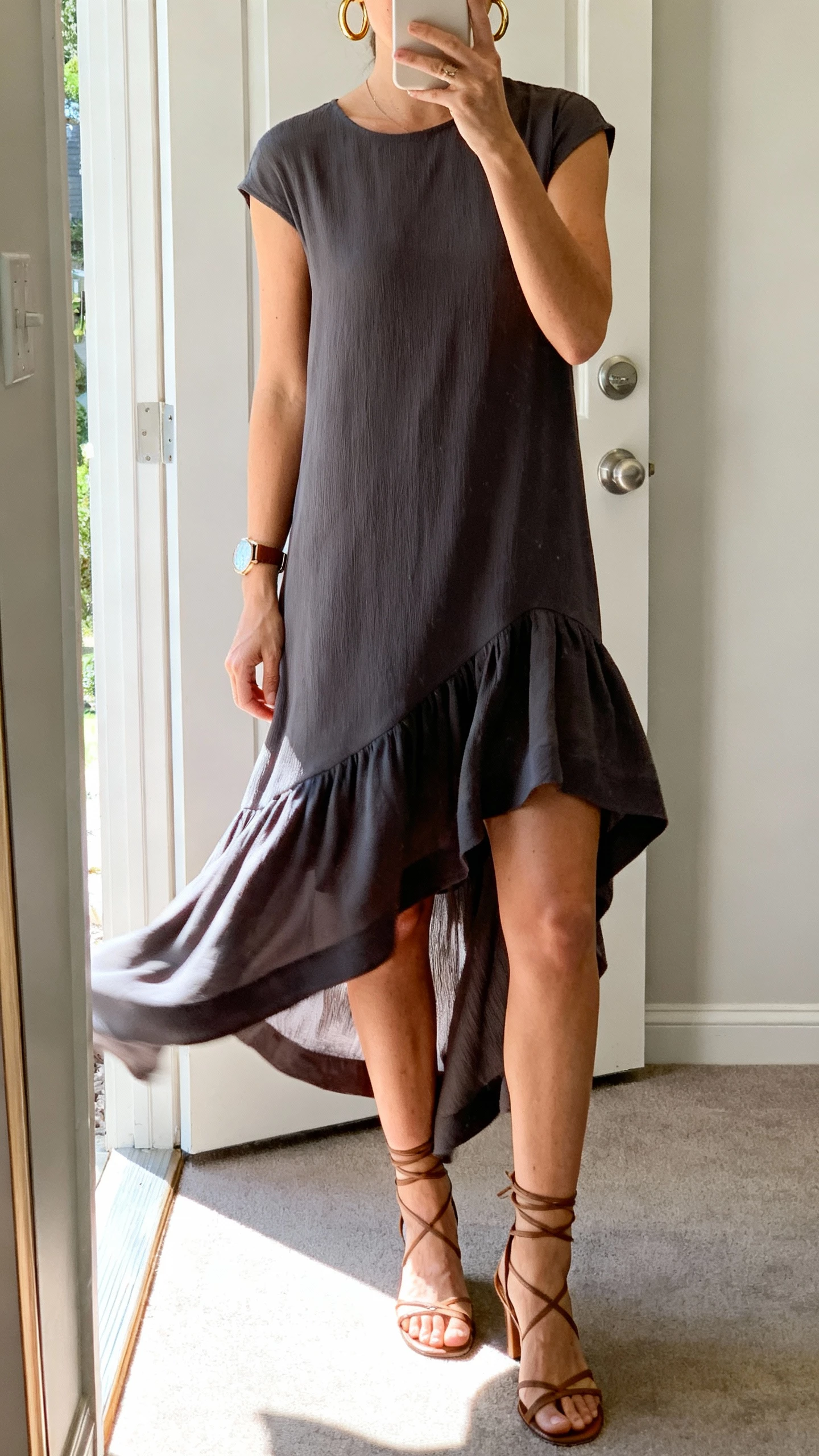 iPhone mirror selfie of a woman wearing a high-low hem dress with flowy movement, windswept-friendly, minimal gold hoop earrings, wristwatch, strappy heels, face not visible, entryway mirror with breeze from open door, afternoon natural light, casual iPhone aesthetic.