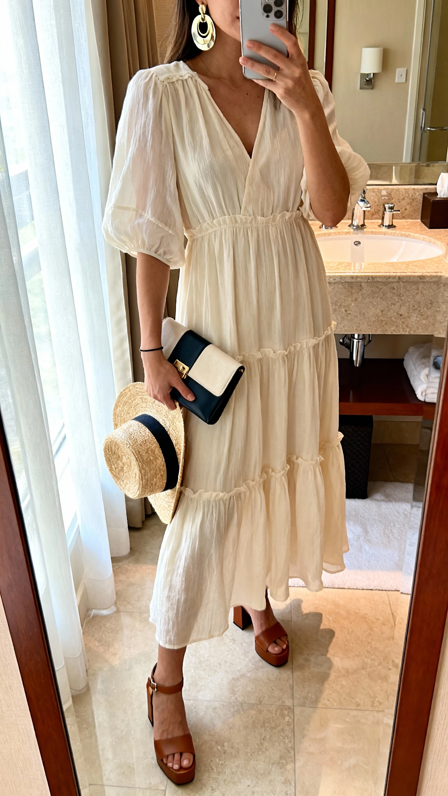 iPhone mirror selfie of a woman wearing a lightweight tiered maxi sundress with block-heel sandals, bold metallic statement earrings, compact contrasting clutch, simple thin cuff, optional straw hat in hand, face not visible, hotel bathroom mirror, morning window light, natural iPhone photo.