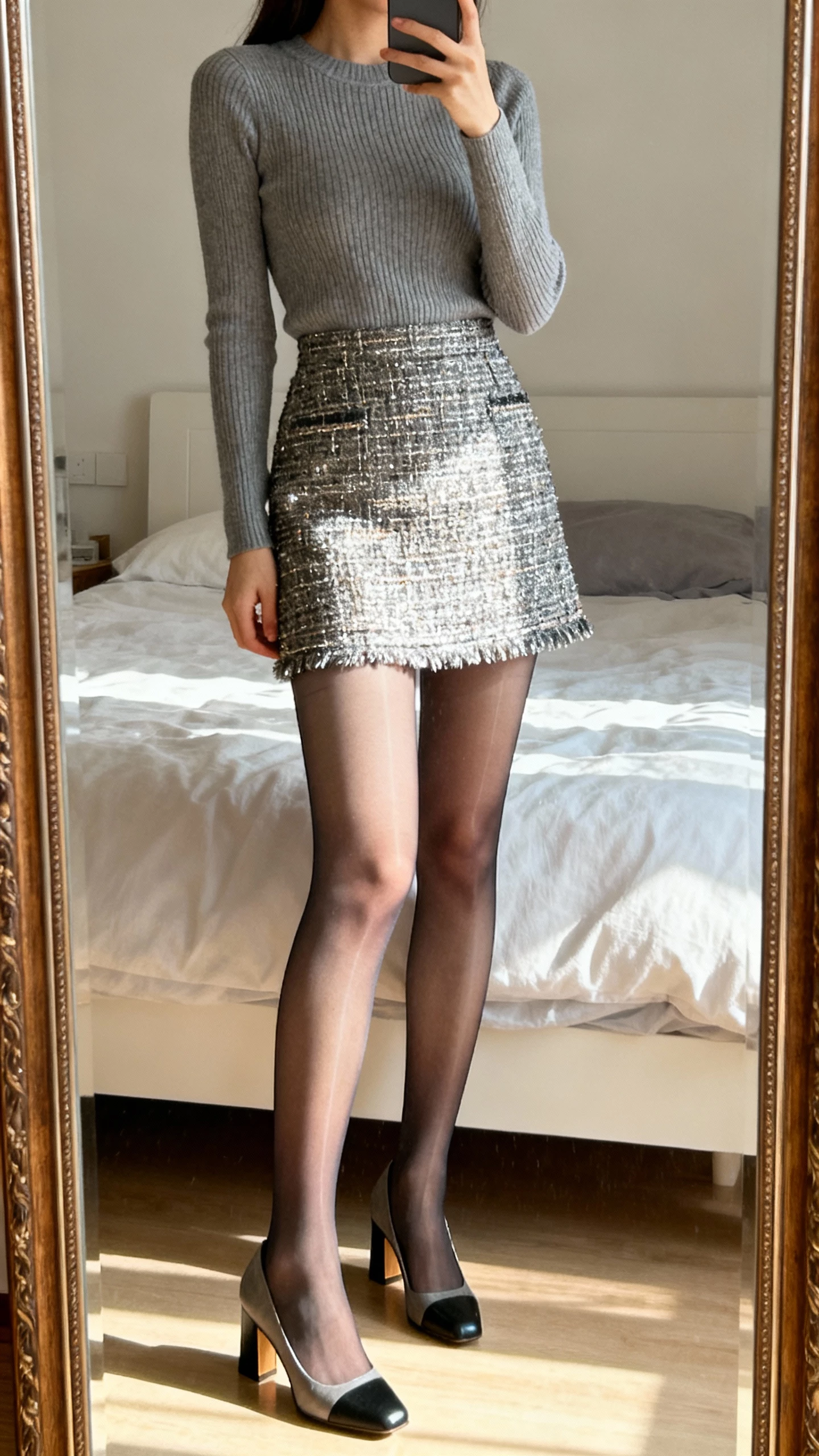 iPhone mirror selfie of a woman wearing a metallic-thread tweed mini skirt with sheer tights, fitted knit top, block-heel pumps, face not visible, cropped at neck, bedroom mirror, morning light, casual iPhone photo