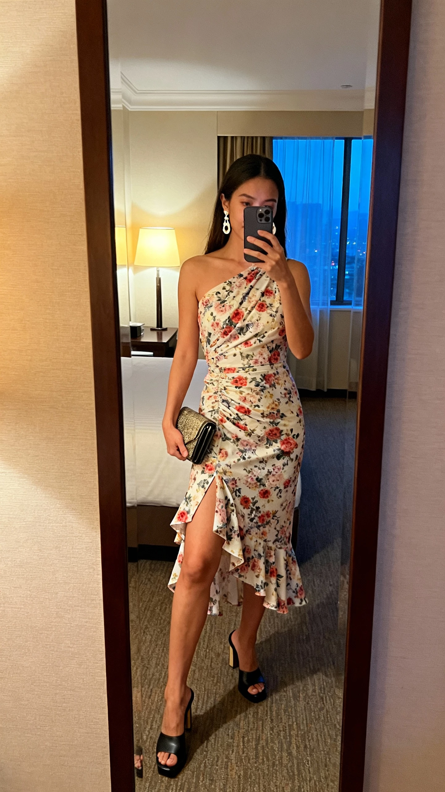 iPhone mirror selfie of a woman wearing a one-shoulder floral midi dress with an asymmetrical hem, heeled mules, drop earrings, and a clutch, face not visible, hotel room mirror, evening lamp and window light mix, natural iPhone photo.