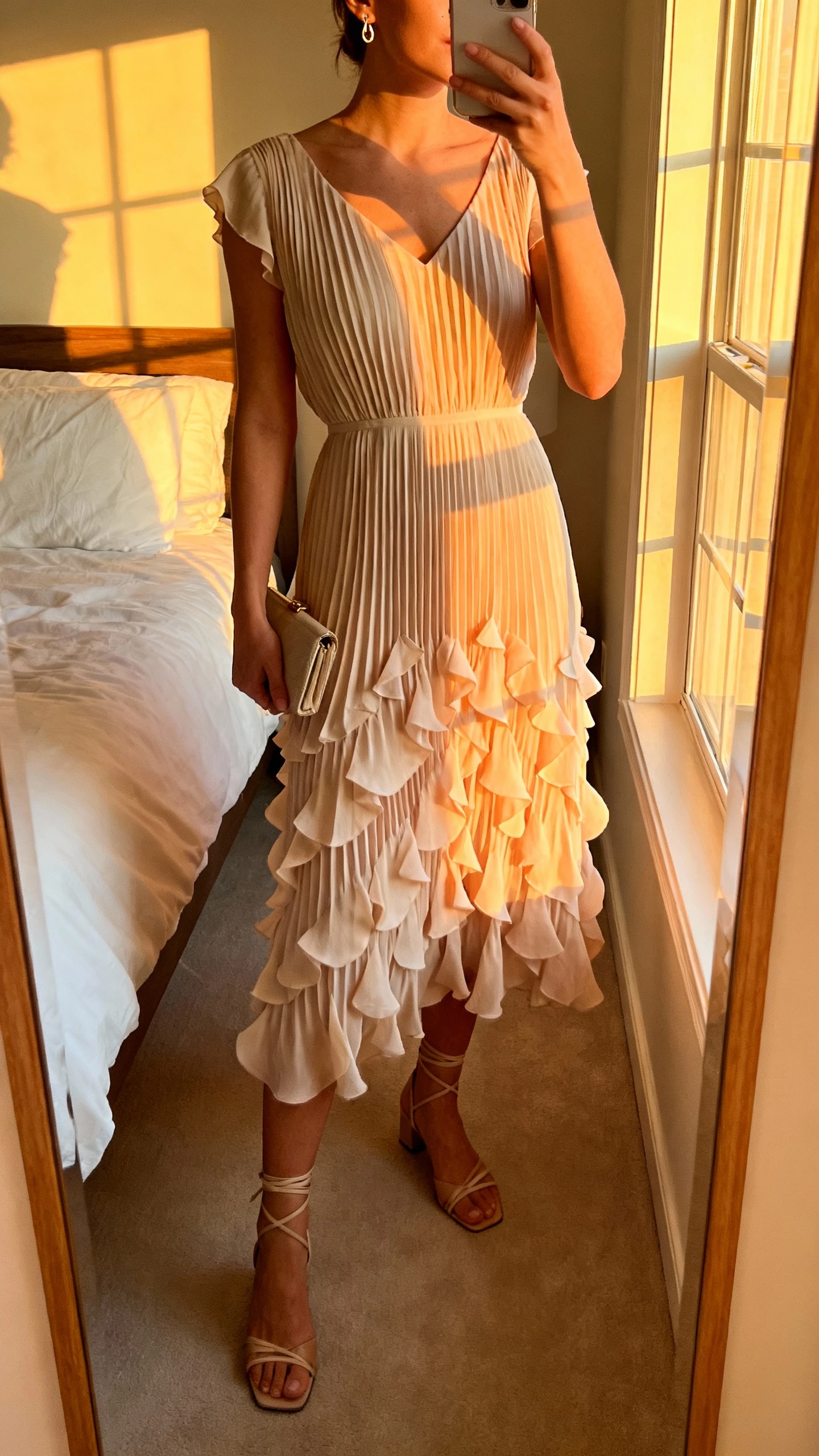 iPhone mirror selfie of a woman wearing a petal-pleated midi dress with delicate flowy pleats, strappy heels, small clutch, dainty earrings, face not visible, cropped at neck, bedroom mirror at golden hour with warm window light, natural iPhone photo quality