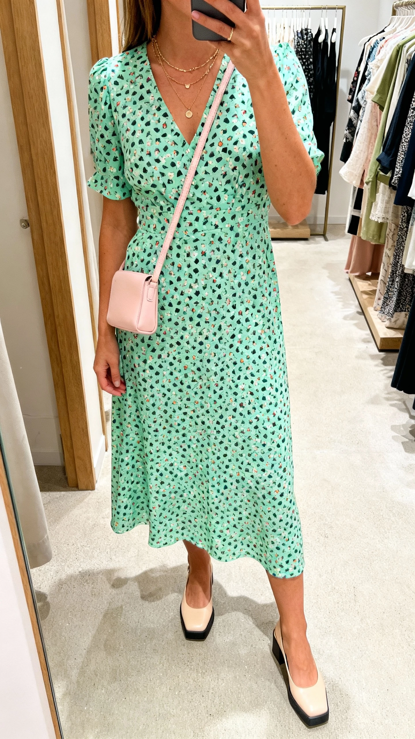iPhone mirror selfie of a woman wearing a pistachio printed midi dress with playful pattern, low block heels, layered delicate necklaces, and a pastel crossbody, face not visible, boutique dressing room mirror, soft overhead lighting, casual iPhone photo.
