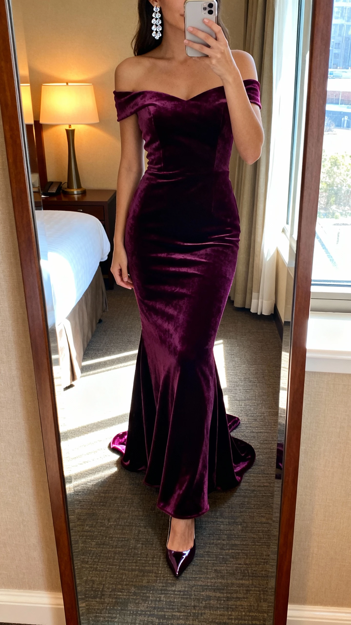 iPhone mirror selfie of a woman wearing a plum velvet off-the-shoulder gown with a mermaid skirt, crystal earrings, and high heels, face not visible, cropped at neck, hotel room full-length mirror, warm lamp light mixed with window light, natural iPhone aesthetic.