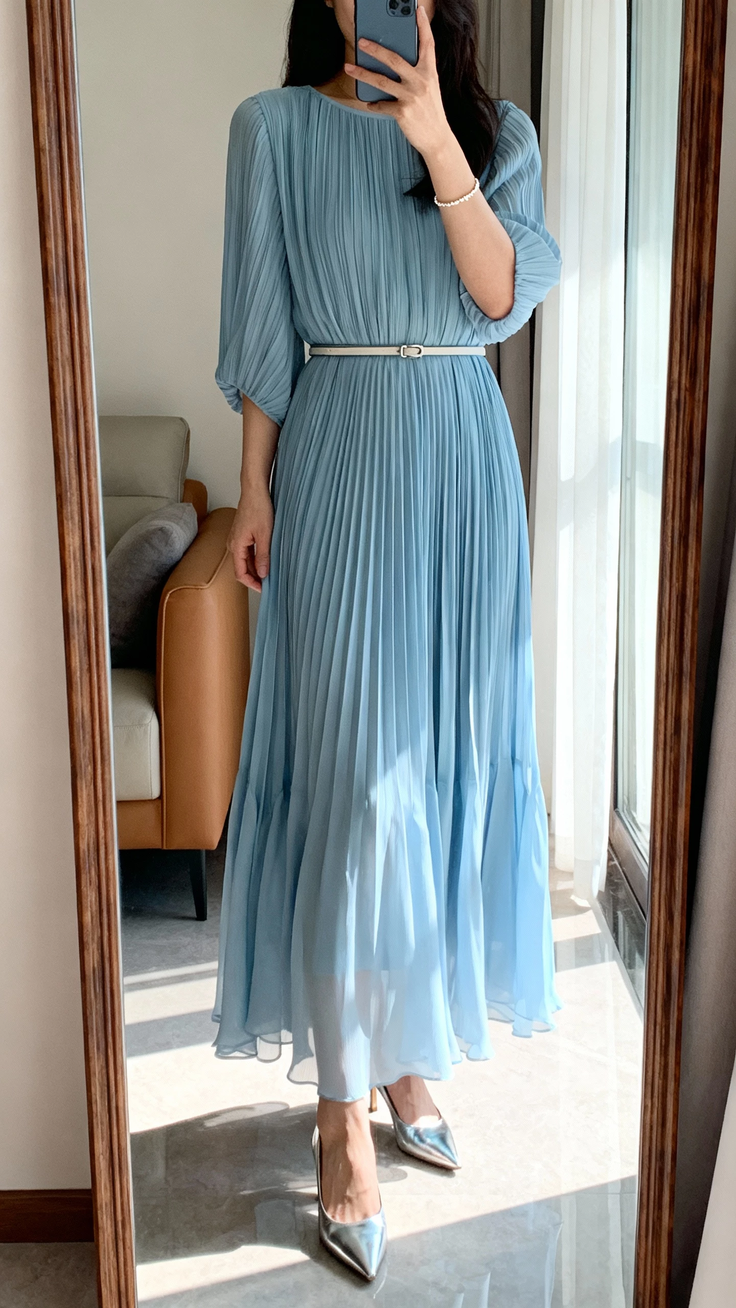 iPhone mirror selfie of a woman wearing a powder blue pleated maxi dress with flowy skirt and thin belt, silver heels, dainty bracelet, face not visible, full-length mirror in a living room with morning window light, natural iPhone aesthetic.