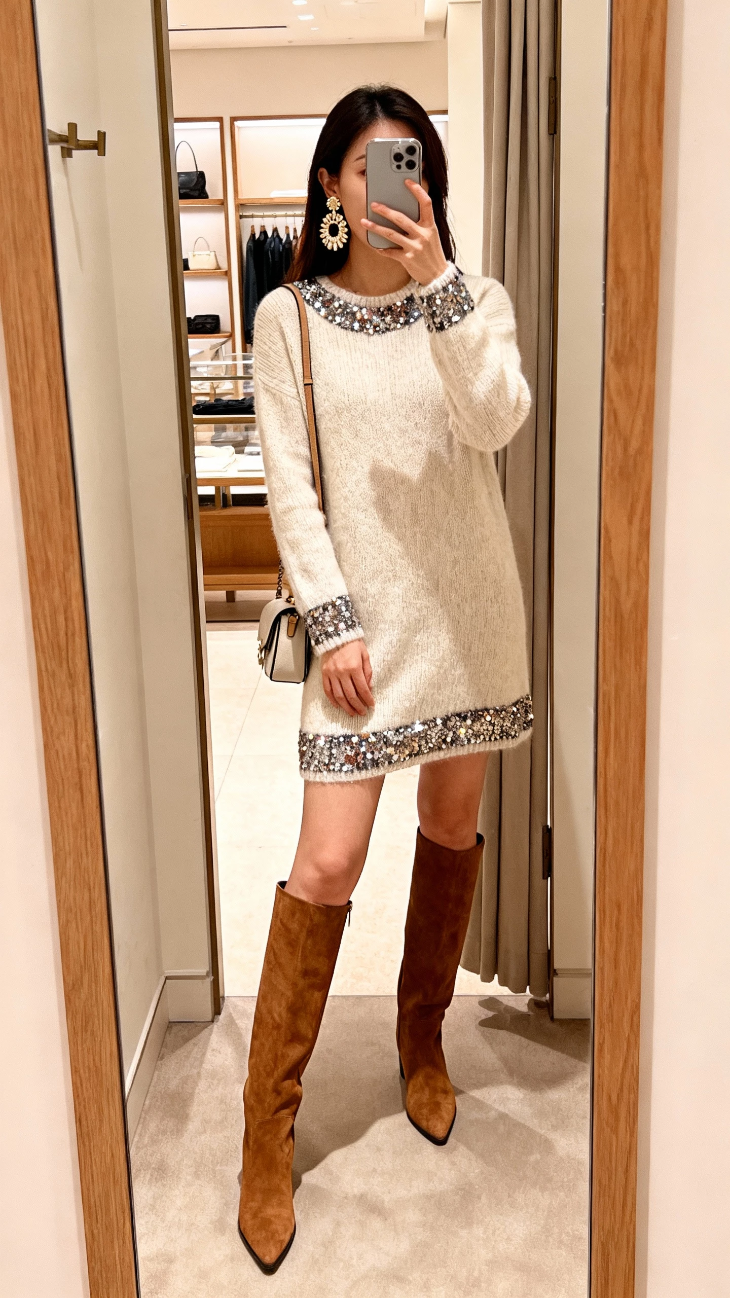 iPhone mirror selfie of a woman wearing a sequin-trim knit dress, statement earrings, and knee-high boots, small shoulder bag, face not visible, boutique fitting-room mirror, warm indoor light, iPhone photo.