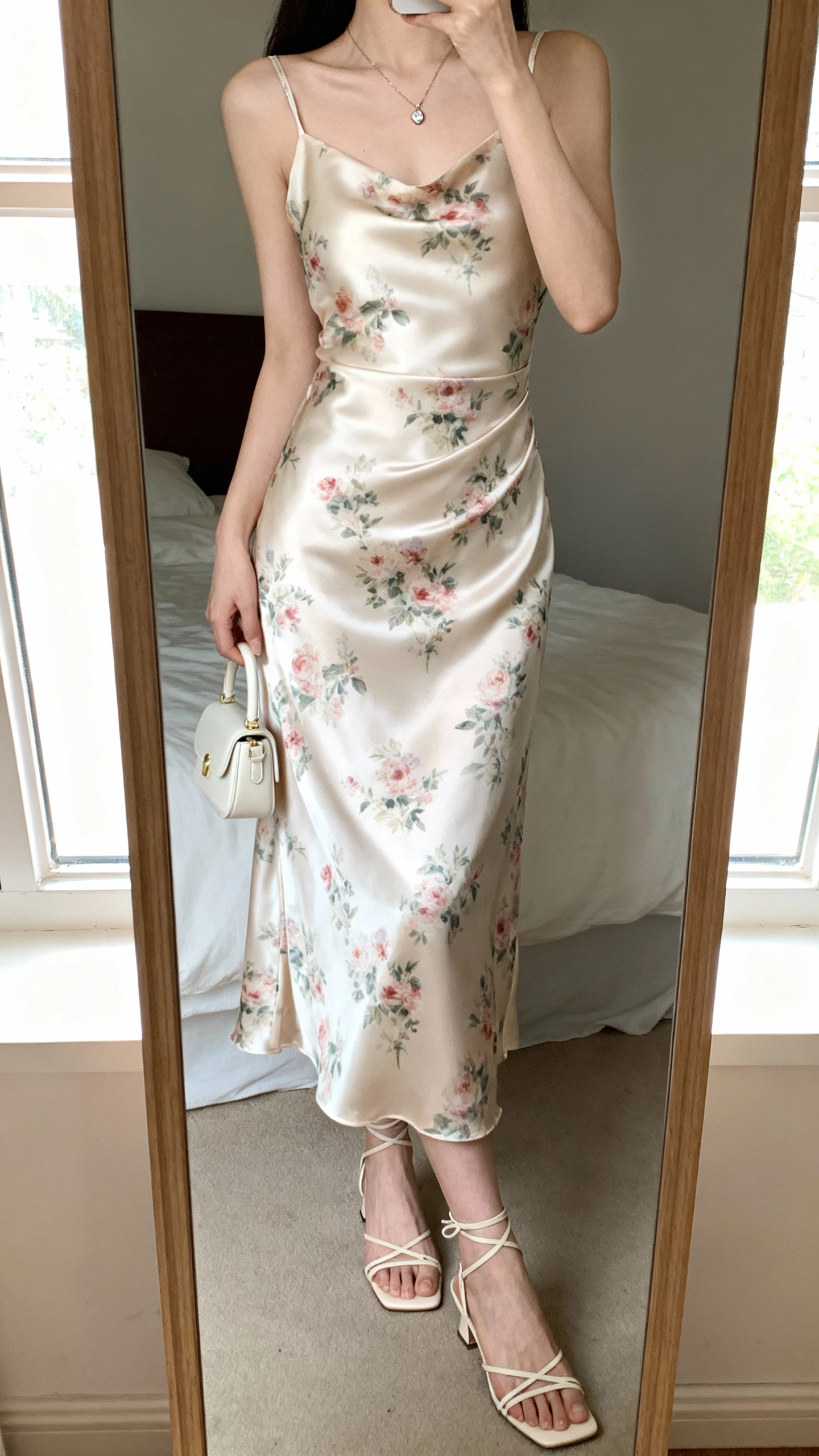 iPhone mirror selfie of a woman wearing a silky garden-party slip dress with a romantic twist (bias-cut slip, soft floral print, delicate strappy heels, dainty pendant necklace, tiny top-handle bag), face not visible, cropped at neck, bedroom mirror with window light, natural iPhone photo quality.