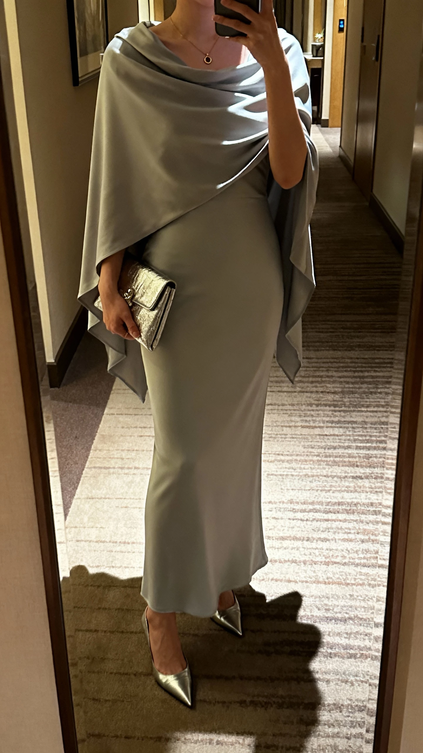 iPhone mirror selfie of a woman wearing a sleek column gown with a draped capelet, pointed heels, metallic clutch, subtle pendant, face not visible, cropped at shoulders in a hotel hallway mirror, evening indoor light, natural iPhone photo quality