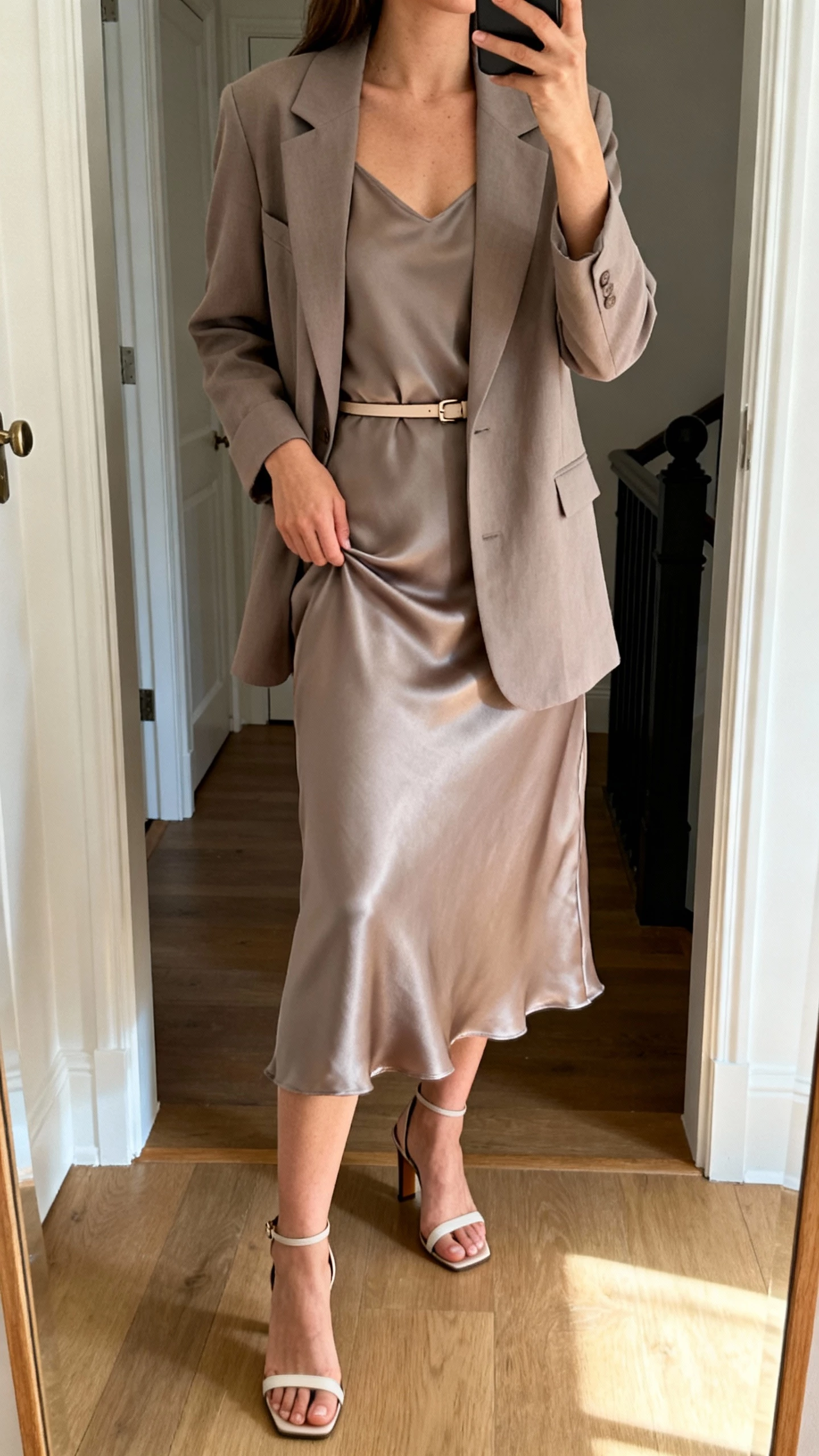 iPhone mirror selfie of a woman wearing a soft taupe blazer over a matching silk slip dress, thin belt, heeled sandals, face not visible, cropped at neck, entryway mirror, natural morning light, iPhone photo quality