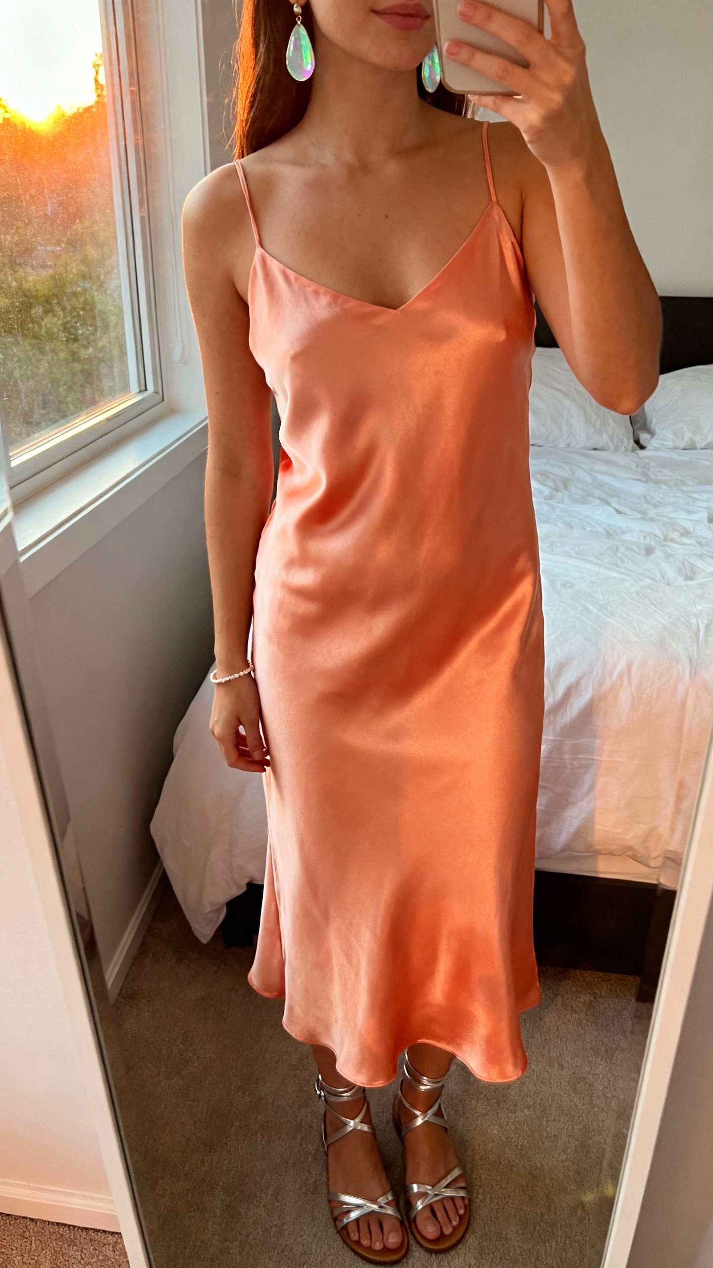 iPhone mirror selfie of a woman wearing a sunset-sorbet satin slip dress with sea-glass shimmer jewelry (iridescent drop earrings and a delicate bracelet), strappy metallic sandals, face not visible, cropped at neck, bedroom mirror with warm sunset window light, natural iPhone photo quality.