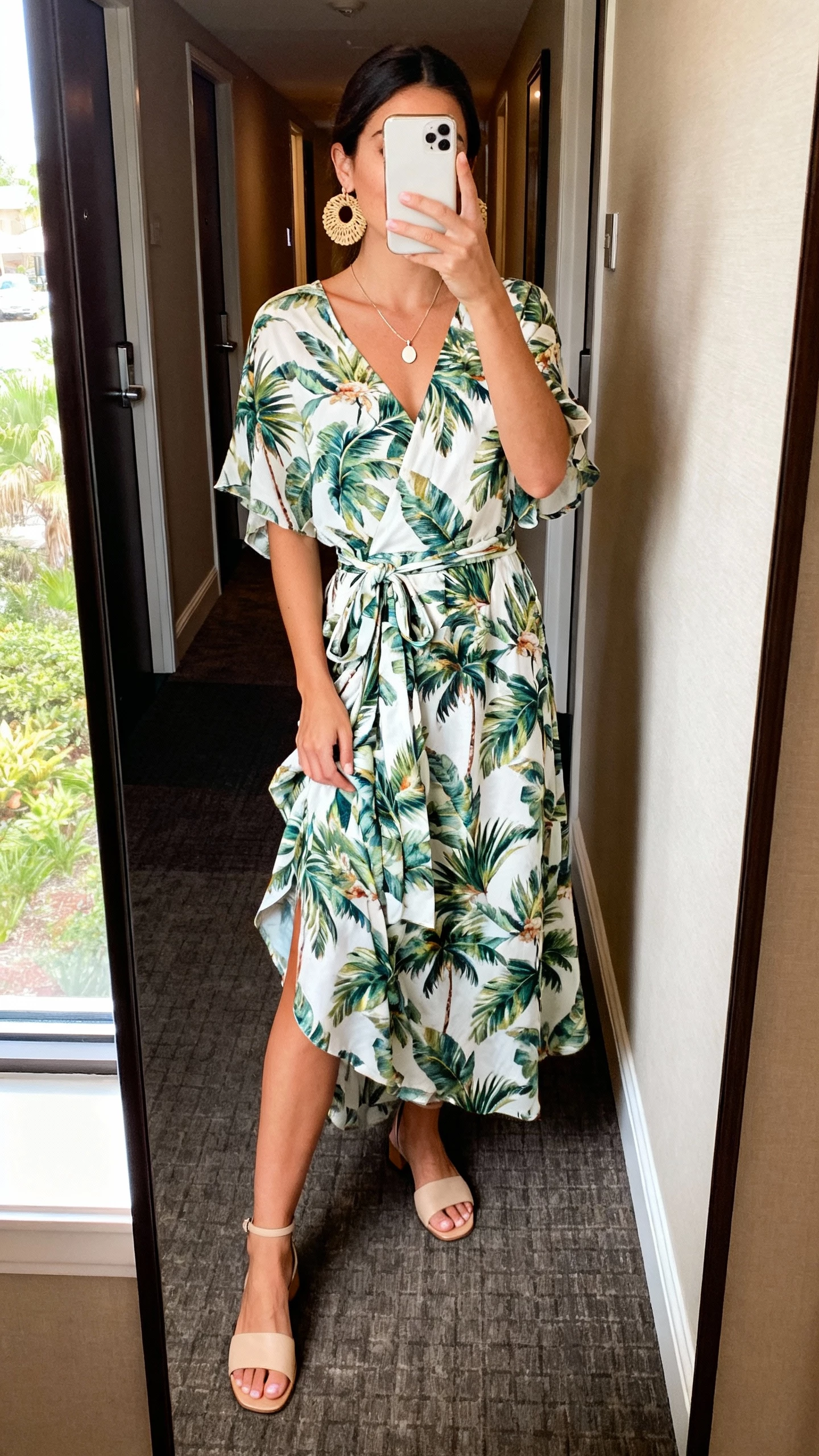 iPhone mirror selfie of a woman wearing a tropical print wrap dress that flutters, delicate pendant necklace, rattan earrings, simple heeled sandals, face not visible, hotel hallway full-length mirror, natural daylight from nearby window, realistic iPhone photo.