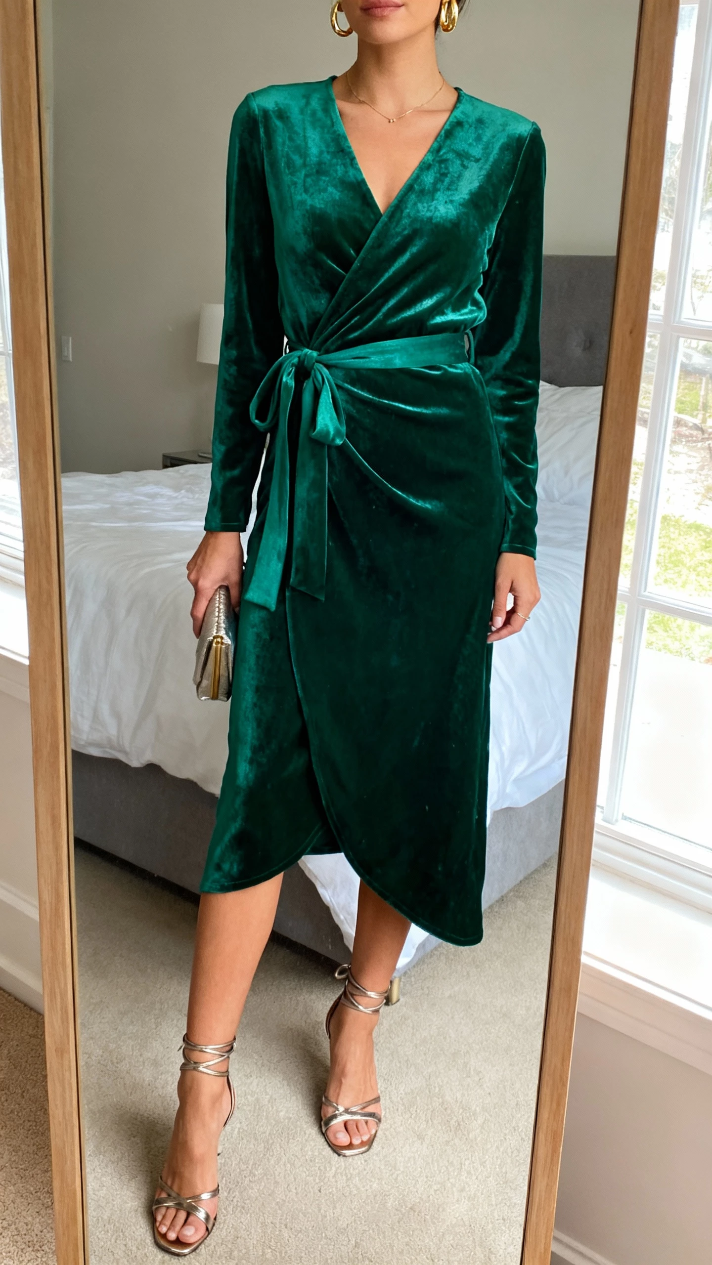 iPhone mirror selfie of a woman wearing an emerald green velvet wrap dress with a tie waist, strappy metallic heels, delicate gold earrings, and a small clutch, face not visible, cropped at neck, bedroom full-length mirror, soft window light, natural iPhone photo quality.