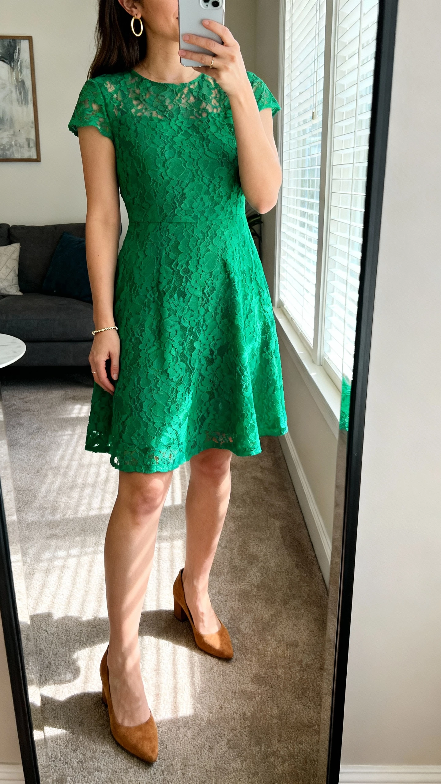 iPhone mirror selfie of a woman wearing an emerald green lace fit-and-flare dress, tan suede pumps, minimal gold hoop earrings, and a dainty bracelet; face not visible, cropped at shoulders, living room full-length mirror, afternoon window light, natural iPhone photo.