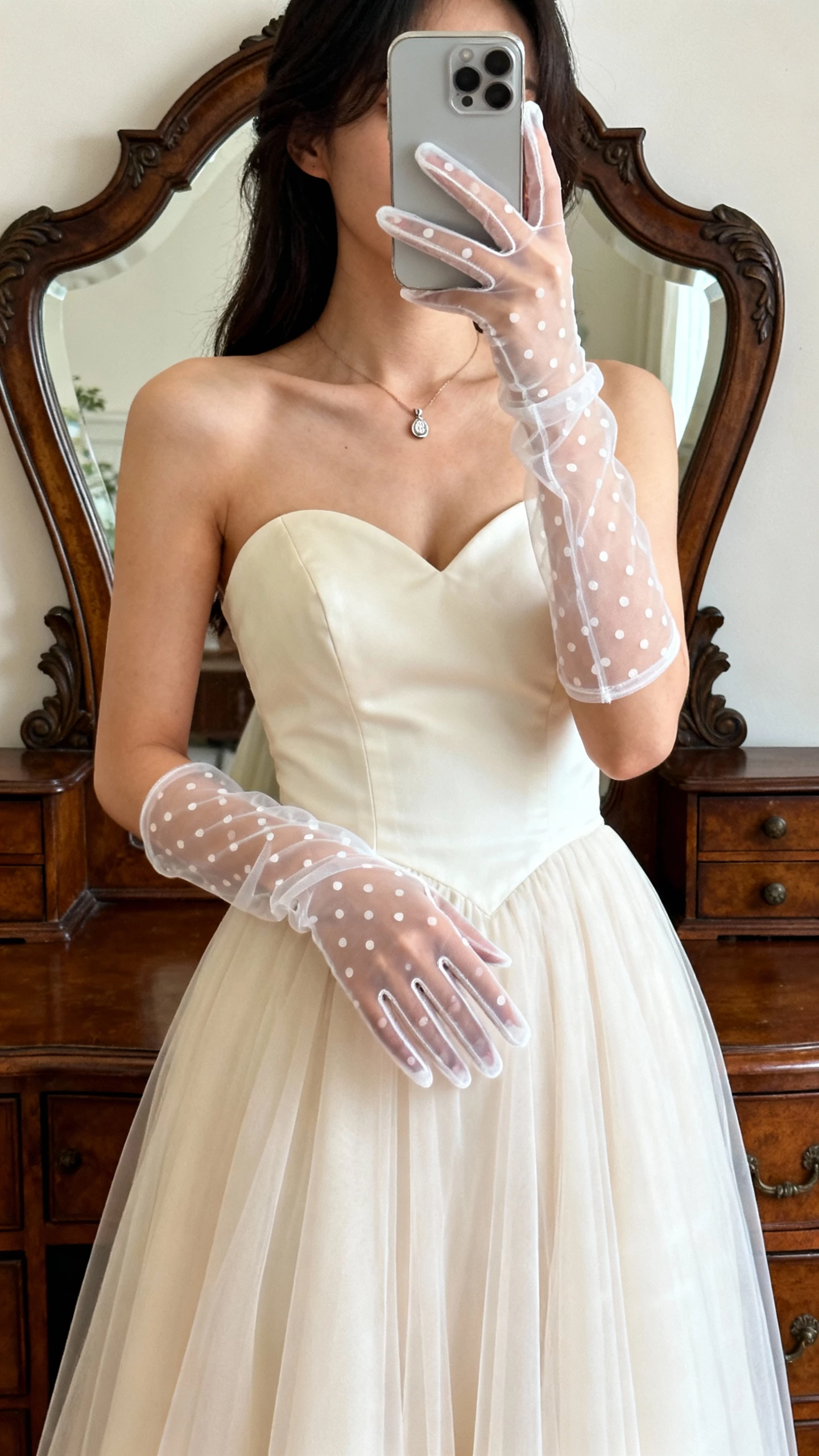 iPhone mirror selfie of a woman wearing illusion dot tulle gloves with a classic sweetheart gown, subtle pendant, face not visible, vintage dresser mirror, soft daylight, natural iPhone photo quality